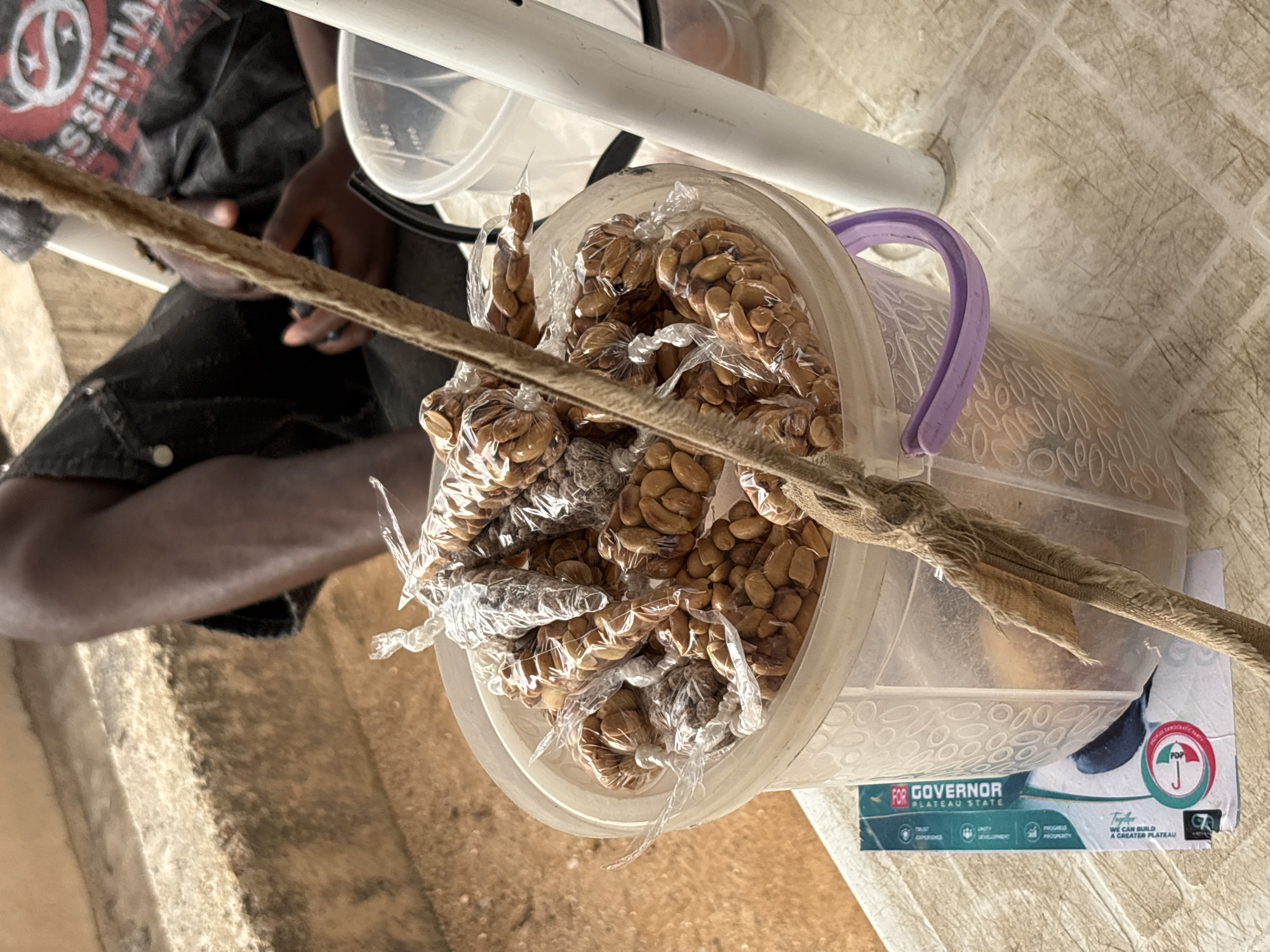 This media file shows a close-up, top-down view of several small bags of roasted peanuts, likely a snack, contained within a clear plastic bucket with a purple handle. The peanuts are golden brown and appear to be individually bagged in clear plastic. A thick, rough rope or strap runs diagonally across the foreground, obscuring some of the peanuts.
In the background, a person's arm and hand are visible, wearing a dark t-shirt with white lettering that partially reads "SSENTIAL". The person appears to be holding a dark object, possibly a phone, and has a gold bracelet on their wrist. The setting suggests an outdoor market or roadside stall, indicated by the tiled floor and a partially visible poster or flyer at the bottom right.
The visible text on the flyer includes "FOR GOVERNOR", "PLATEAU STATE", and other phrases like "TRUST", "EXPERIENCE", "UNITY DEVELOPMENT", and "PROGRESS PROSPERITY". There is also a logo with an umbrella symbol and text that includes "DEMOCRATIC PARTY" and "Together WE CAN BUILD A GREATER PLATEAU". This suggests the photograph was taken in Plateau State, Nigeria, likely in Jos, and the context relates to a political campaign or event. The lighting suggests it is daytime.