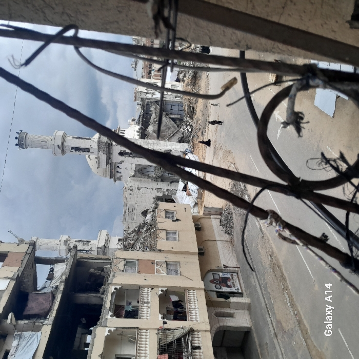 An aerial perspective photograph captured in Khan Yunis, Palestinian Territory, depicts a street scene characterized by extensive structural damage under an overcast sky. In the immediate foreground, blurred dark metal bars and wires, possibly from a damaged railing, partially obscure the view. To the left, a multi-story residential building exhibits severe damage, with large sections of its facade missing, exposing interior rooms and damaged balconies where laundry hangs. Debris is scattered at its base.
Across a relatively clear asphalt street, several individuals are walking. Two prominent figures are visible: one person dressed in a black abaya walks on the left side of the street, and another individual is further to their right, accompanied by a smaller figure, possibly a child. More distant figures are faintly visible further down the street. The street itself appears largely intact but leads into areas strewn with rubble and collapsed building materials.
In the background, a large, distinctive building, identified as a mosque, dominates the horizon. It features a central dome and two minarets, both of which show significant damage. The mosque's main structure appears heavily compromised, with large sections of its walls collapsed and debris surrounding its base. The overall environment indicates widespread destruction of urban infrastructure.
