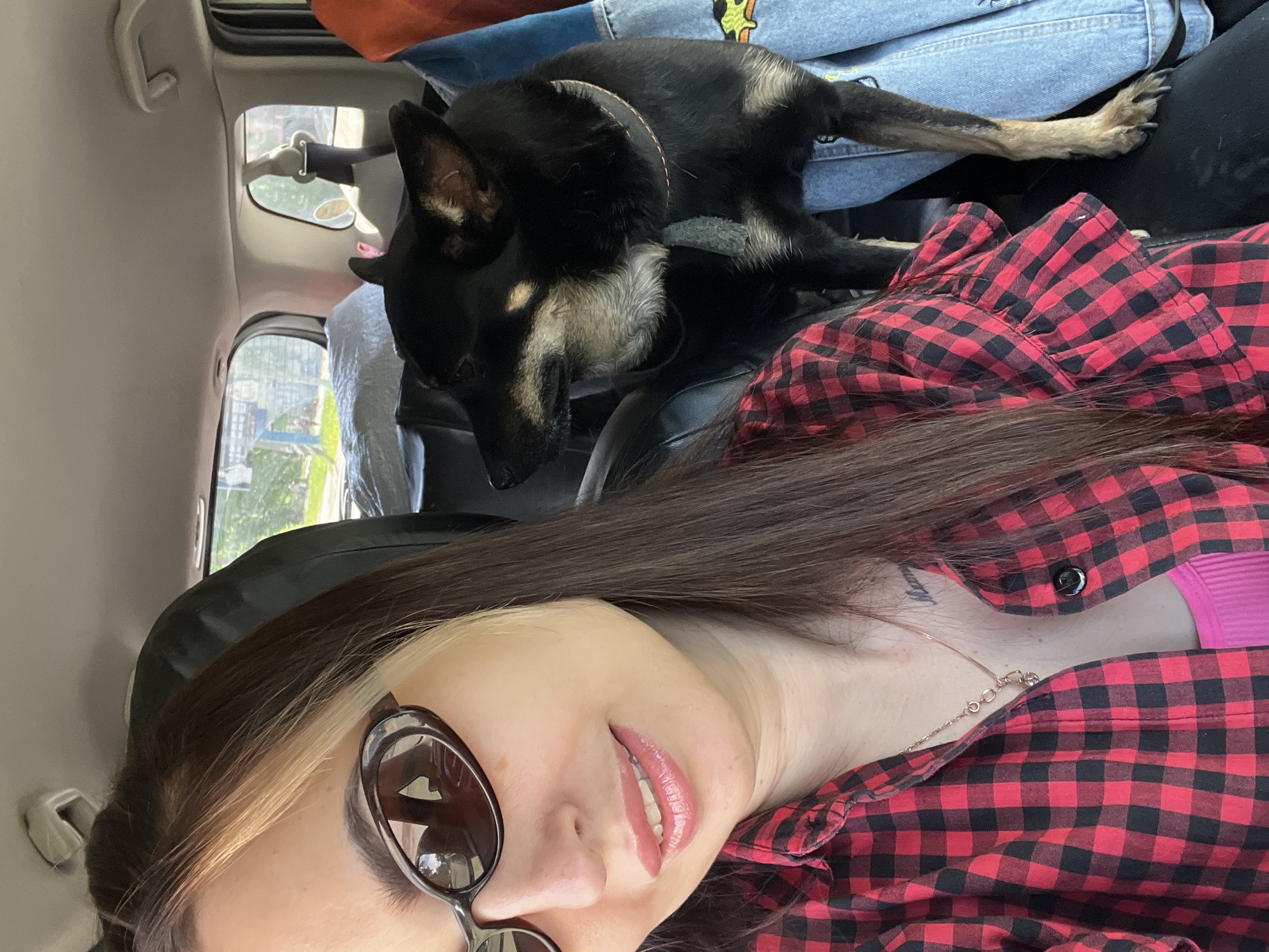 A woman is taking a selfie inside a vehicle, with a dog resting behind her. The photo is taken from a slightly upward angle, showing the woman's face and upper torso in the foreground, with the dog and vehicle interior in the background.
The woman has long, dark hair with a distinct blonde streak near her face. She is wearing dark, cat-eye style sunglasses, a red and black plaid shirt, and a gold-colored chain necklace. A hint of a pink top is visible underneath her plaid shirt, and she has light pink glossy lipstick. A faint tattoo with indistinct text is visible on the left side of her neck/shoulder. Her expression is a relaxed slight smile.
Behind the woman, a black and tan dog, possibly a German Shepherd mix, is resting or sleeping. The dog has black fur with tan markings on its face, chest, and legs, and is wearing a simple tan collar. It appears to be lying on some light blue denim fabric (possibly jeans) and other items, including an orange bag, in the back of the vehicle. One of the dog's front paws with visible claws is resting on the denim.
The interior of the vehicle has a light-colored ceiling. Through the rear windows, greenery and distant structures or buildings are visible, suggesting an outdoor setting during the daytime. The bright lighting implies it is sunny. A grab handle is visible on the car's interior ceiling. The overall atmosphere appears casual and relaxed, perhaps during a road trip or outing.