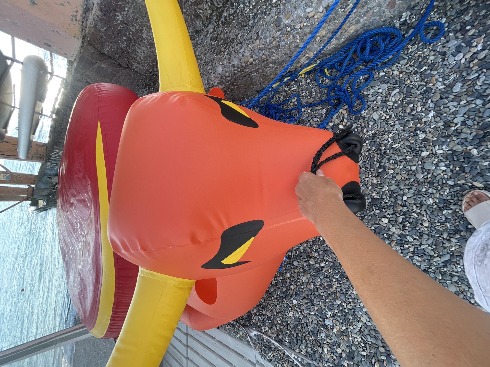 In Sochi, Russia, on a pebble beach, an inflatable bull with orange body, yellow horns, and black and yellow eyes is positioned on the ground. A person's hand is holding a black rope attached to the bull's muzzle. Blue ropes lie on the pebbles next to the bull. In the background, a red and yellow inflatable cushion is visible, with the sea and a pier structure beyond it.