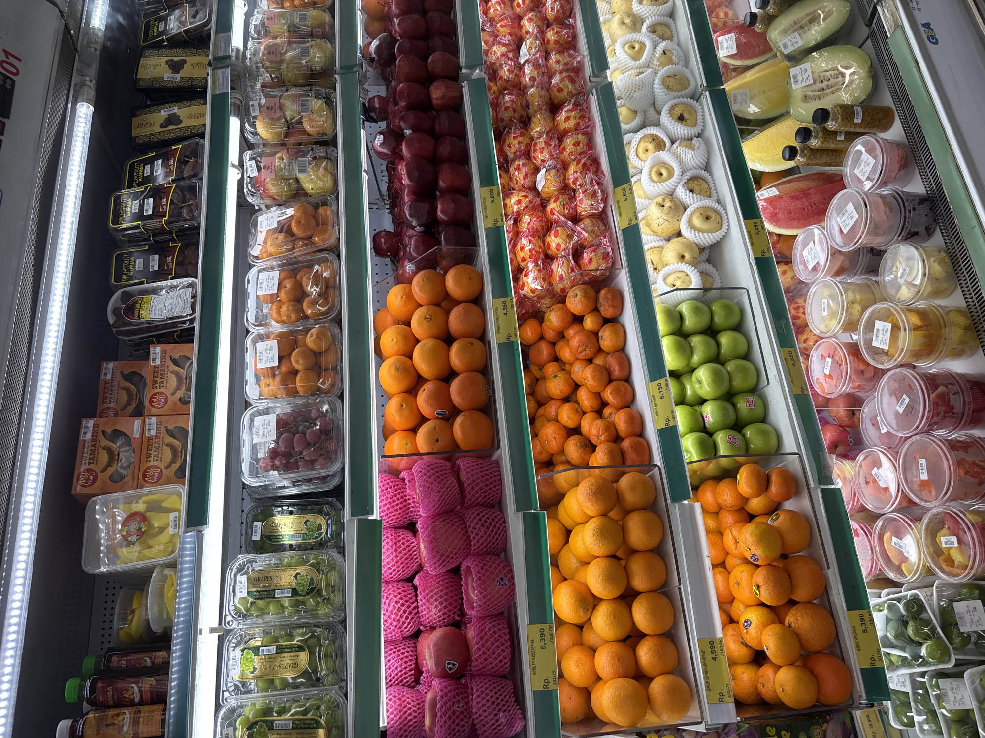 A refrigerated supermarket display case is filled with various fruits and processed fruit products. The scene is likely in Kuta Selatan, Indonesia, given the pricing labels using "Rp." (Rupiah).
Shelves, from left to right, contain: dates and tamarind in boxes and plastic containers; green grapes, mandarin oranges, and small apples in pink foam netting; mandarin oranges; red apples and mandarin oranges; red and yellow apples in clear plastic packaging; Asian pears in white foam nets; green apples; mandarin oranges and limes; and sliced honeydew, watermelon and cut fruit in plastic containers.
Each section has price markers in Indonesian Rupiah.
The lighting is bright and fluorescent, typical of a supermarket.