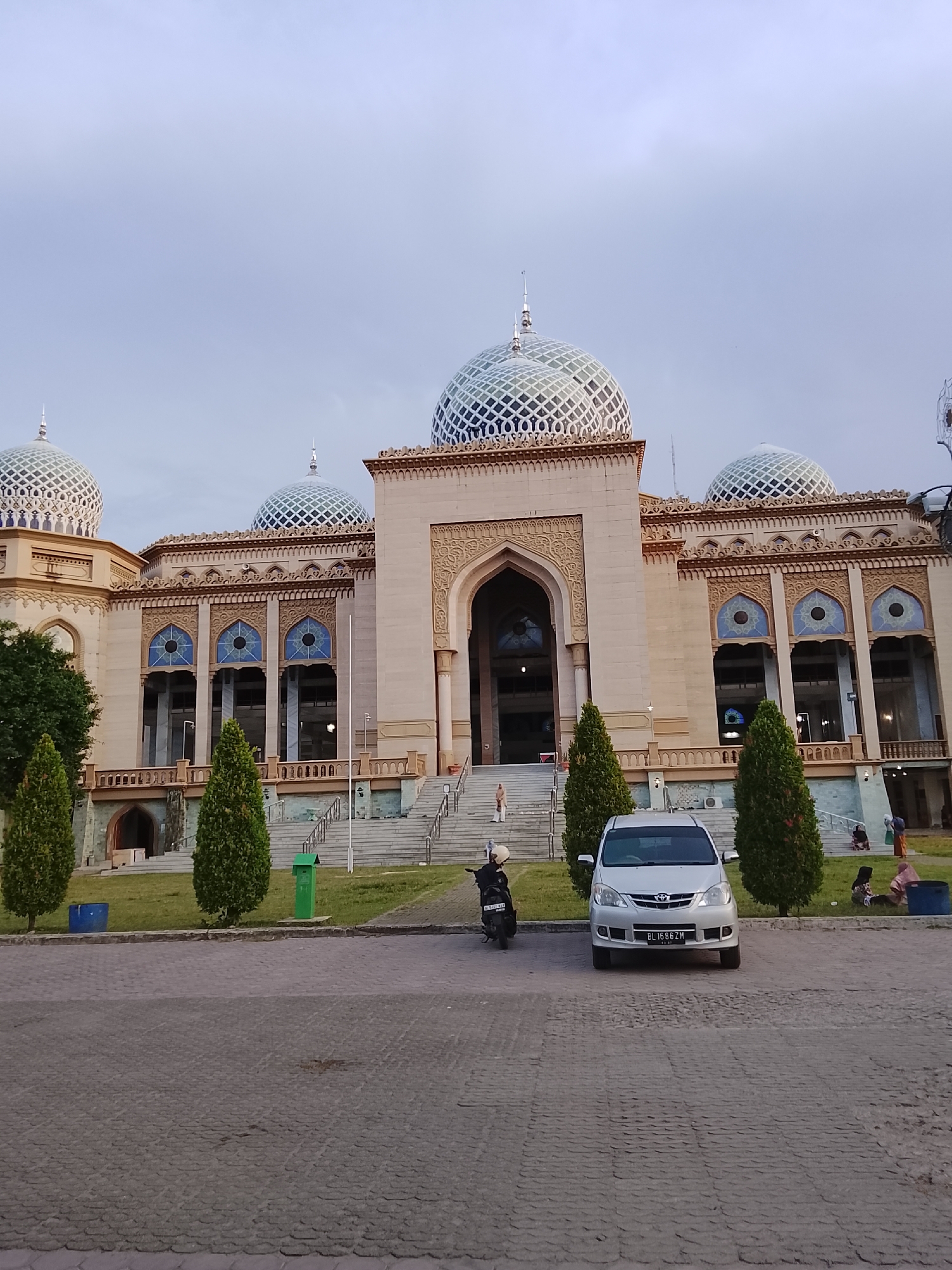 The image captures the exterior of the Islamic Center of Lhokseumawe, Indonesia, an ornate mosque with multiple domes and intricate architectural details. The mosque features a central archway, flanked by large windows with blue stained glass and decorative carvings. The building is constructed with light-colored stone. In front of the mosque, a paved area leads to a grassy lawn. A silver car is parked on the paved area, with a motorcycle positioned to its left. Several cypress trees are planted on the lawn. In the background, on the steps of the mosque, a few people are visible, some sitting and others standing. The sky is overcast and appears to be late afternoon or early evening. The overall scene depicts a peaceful and serene environment outside a prominent religious building.