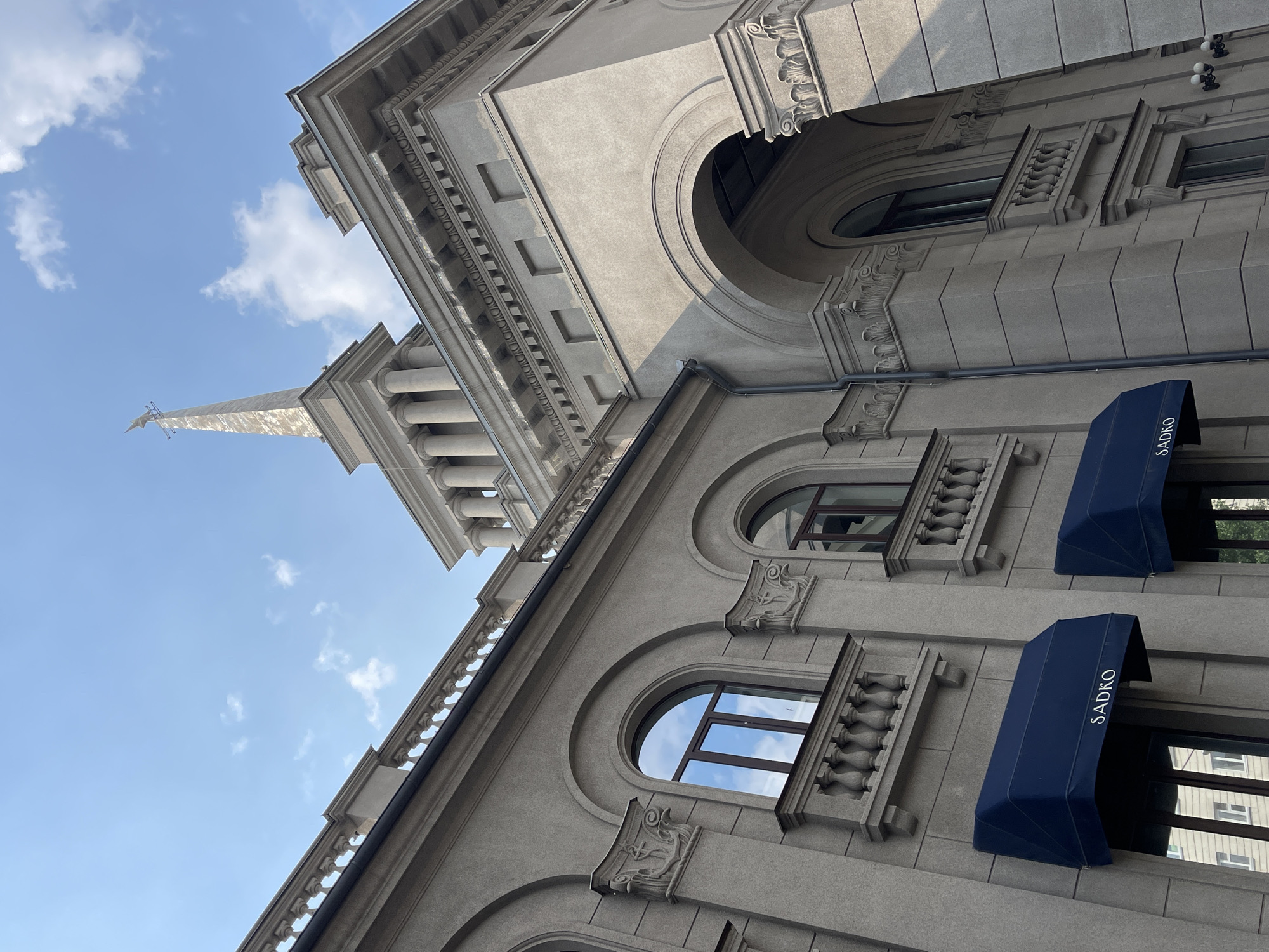 This is a low-angle, exterior, daytime, eye-level shot of a large, light-gray, stone building in Krasnoyarsk, Russia. The building features classical architectural elements such as arches, columns, and decorative moldings. A prominent spire rises from the center of the building, topped with a bird-like weather vane. Two blue awnings with the word "SADKO" are visible on the lower level of the building. The sky above is clear blue with a few white clouds. The scene is static with no people or discernible activity.