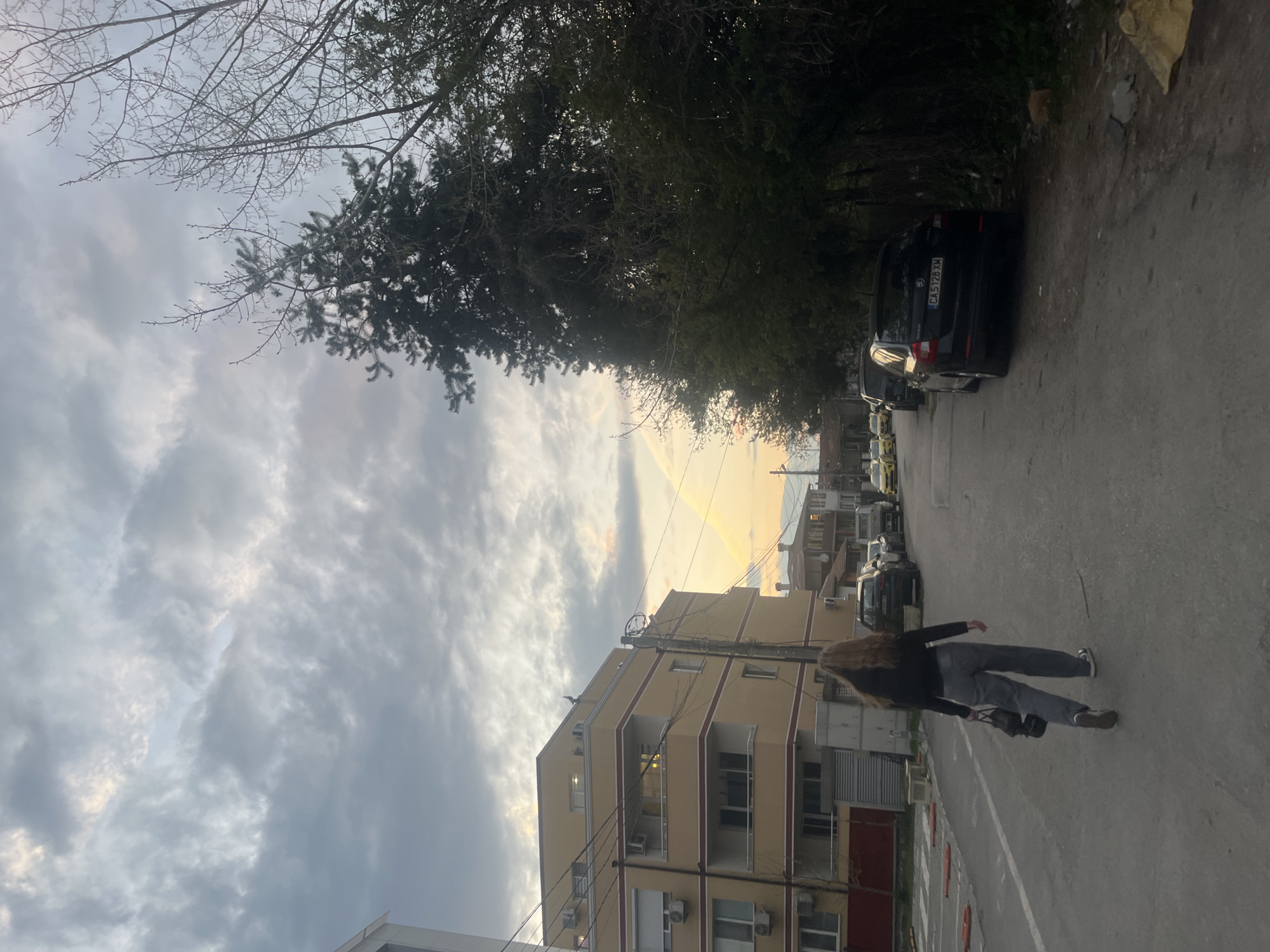 The image displays an outdoor street scene in Sofia, Bulgaria, captured during either sunrise or sunset, evidenced by the soft, golden light breaking through an overcast sky. A person with long, light-brown hair, dressed in a dark jacket and grey pants, walks away from the viewer down the middle of a paved street, carrying a dark shoulder bag.
On the left, a multi-story residential building painted yellow with prominent red trim is visible. It features balconies and several air conditioning units attached to its exterior. Along the right side of the street, several cars are parked, including a black vehicle with the license plate "CA 5128 XM", where "CA" signifies Sofia. A line of dense green trees shades this side of the road, and additional parked cars and what appear to be yellow taxis can be seen further in the distance. Bare tree branches are visible against the cloudy sky in the upper left portion of the image. The sky itself is filled with grey, textured clouds, but a horizontal band of warm, golden light is prominent near the horizon, casting a gentle glow over the scene. The atmosphere suggests a quiet, residential urban setting.