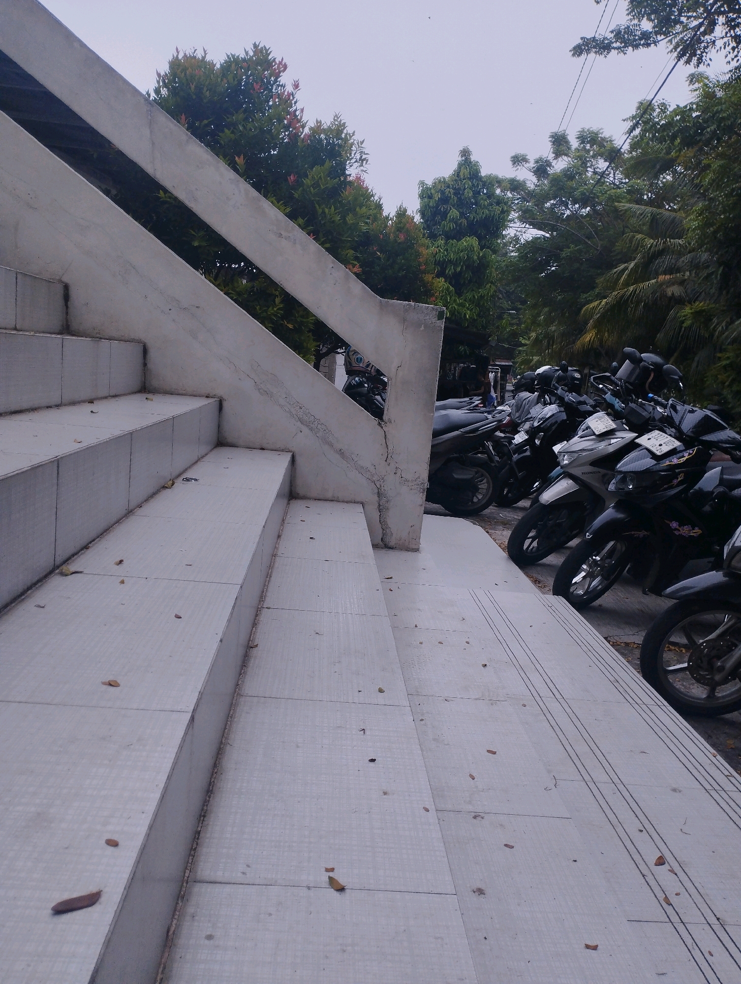 The image depicts an outdoor scene featuring a concrete staircase on the left and a row of parked motorcycles on the right. The staircase is light gray and has several steps, with a concrete railing extending upwards and to the right. Scattered leaves are visible on the steps. Beyond the staircase, a line of motorcycles, mostly scooters, are parked. The motorcycles are varied in color, including black, silver, and white. Some have visible license plates. Behind the motorcycles, lush green trees and vegetation form a backdrop, suggesting an urban or suburban environment. The sky is overcast and a pale gray. The scene appears to be in an open area, possibly a parking lot or a walkway adjacent to a building. The overall impression is one of an everyday scene in a tropical or subtropical climate, with the presence of motorcycles indicating a common mode of transportation. It is plausible that this image was taken in the city of Padang, Indonesia, given the prevalence of motorcycles and the type of vegetation visible.