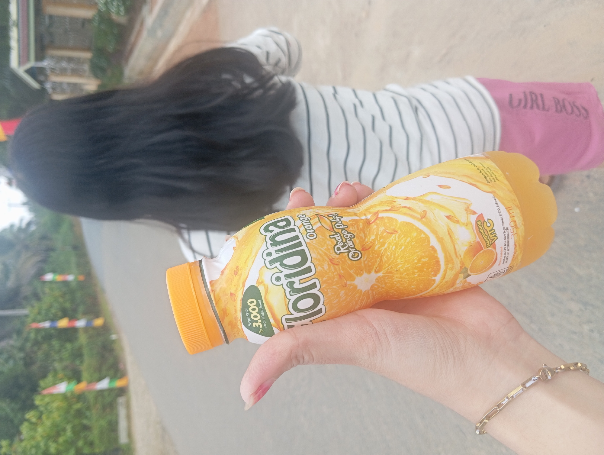 A person's hand, adorned with a metallic bracelet, holds an orange-flavored juice bottle. The bottle's label is primarily yellow and orange, featuring images of sliced oranges and the brand name "Floridina" in a distinctive font. The price, "Rp 3.000", is visible on a green sticker on the label. In the background, a child with long, dark hair walks away from the camera, wearing a horizontally striped t-shirt and pink shorts with the text "GIRL BOSS" printed on them. The scene appears to be outdoors, with a paved road and greenery visible. Three Indonesian flags on poles are positioned along the side of the road, suggesting the location is Rejosari, Indonesia, possibly during a local celebration or event.