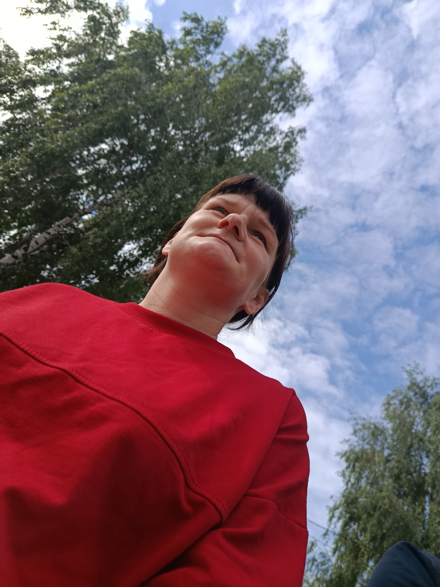 The image, captured in the city of Фоки, Russia, presents an individual from an extreme low-angle perspective, facing upwards and slightly to their right. The person, wearing a solid red long-sleeved top, occupies the lower portion of the frame, with their head and shoulders prominently featured. Dark, short hair with bangs frames a face that exhibits a concentrated upward gaze, characterized by a slightly furrowed brow and an elevated chin. The background consists of a bright blue sky with scattered white clouds on the right side of the frame, and dense green deciduous tree canopies, including visible lighter-colored tree trunks, on the left. The entire scene is illuminated by natural daylight.