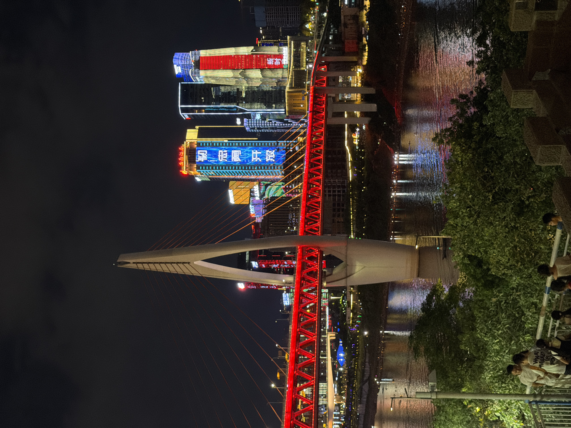 The image captures a nighttime cityscape in Yuzhong District, China, featuring a prominently illuminated red bridge spanning across a river. The bridge's structure is intricate, with a central white pylon supporting red cables. On the far bank of the river, several tall buildings are adorned with colorful, bright lights, including vertical text displays in Chinese characters, predominantly in blue and red. The water of the river reflects the city's lights, creating a shimmering effect. In the foreground, there is dense greenery and a few people standing on a raised platform, some looking towards the illuminated bridge and buildings. A streetlamp is visible to the left of the frame. The sky is dark, indicating it is night.
