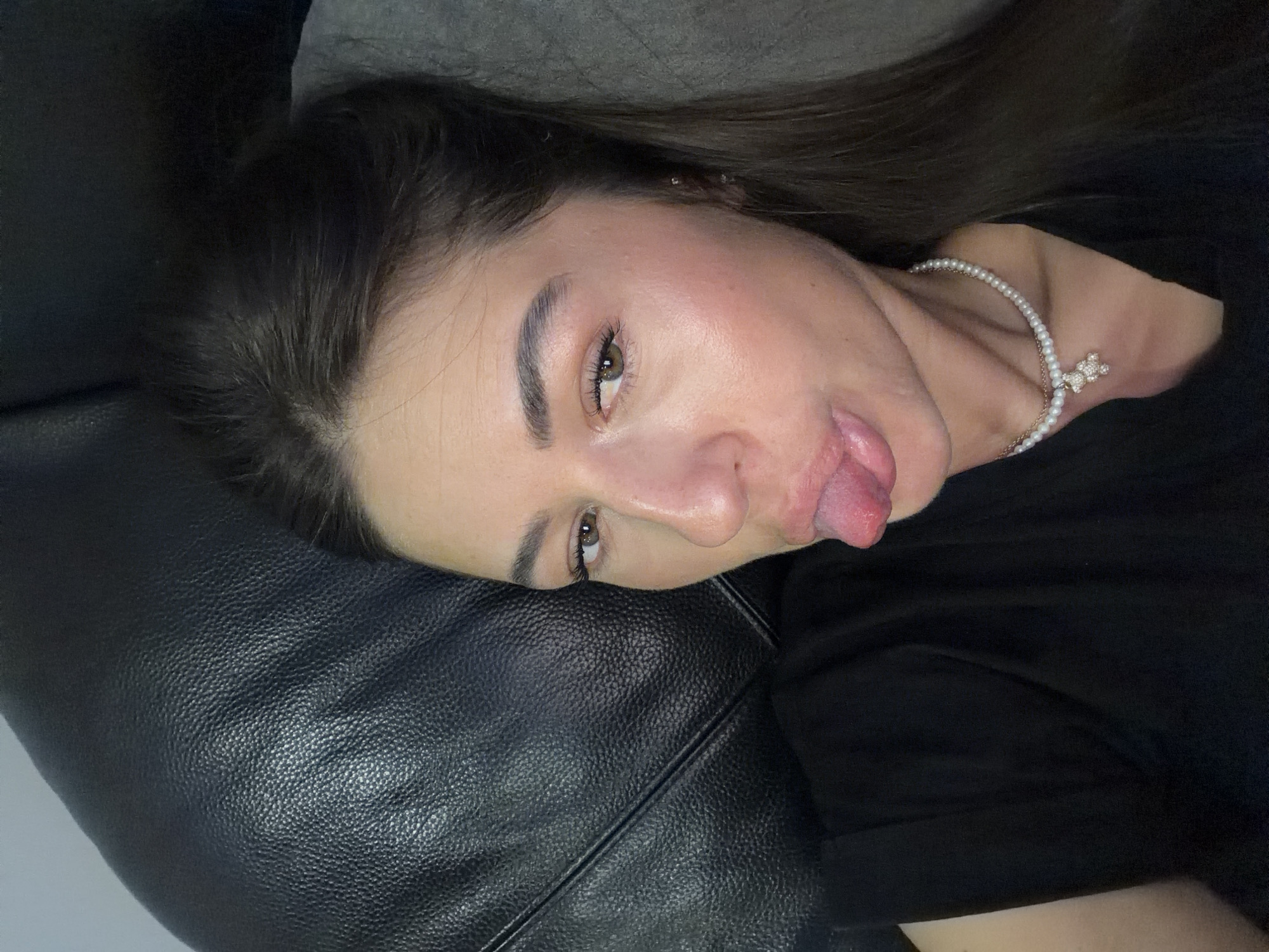 A young woman with dark hair is lying on a black, textured couch, possibly a sofa or a headboard, in a dimly lit room. She is wearing a black t-shirt and a pearl necklace with a sparkling pendant shaped like a teddy bear. Her right eye is visible, and she is sticking her tongue out, giving a playful or cheeky expression. The background suggests an indoor setting, with a gray surface to her right, possibly another piece of furniture or a wall. The lighting highlights the sheen on her skin and the texture of the couch. There is no indication of the city of Novi Iskar, Bulgaria, or any specific geographical markers within the image.