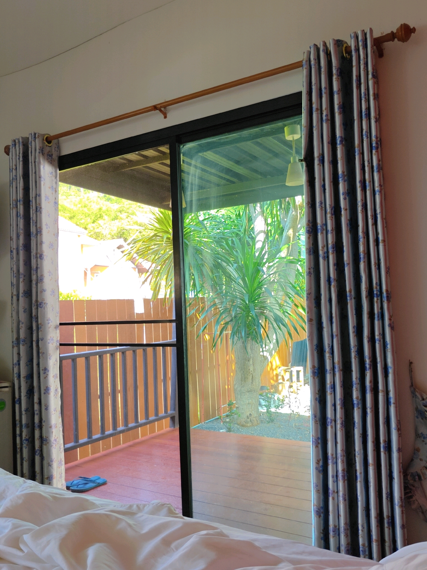 The image depicts a bedroom interior with a view of an outdoor patio area through a sliding glass door. A wooden curtain rod spans the top of the door frame, from which two patterned curtains are hung. One curtain is partially drawn open to the left, revealing the outdoor scene. The outdoor area features a wooden deck with a railing, a large potted plant with palm-like leaves, and a wooden fence in the background. Sunlight illuminates the outdoor space. Inside the bedroom, white bedding is visible in the foreground. A pair of blue flip-flops rests on the outdoor deck. The overall setting suggests a tropical or subtropical environment. The location is identified as Khao Yai, Thailand.