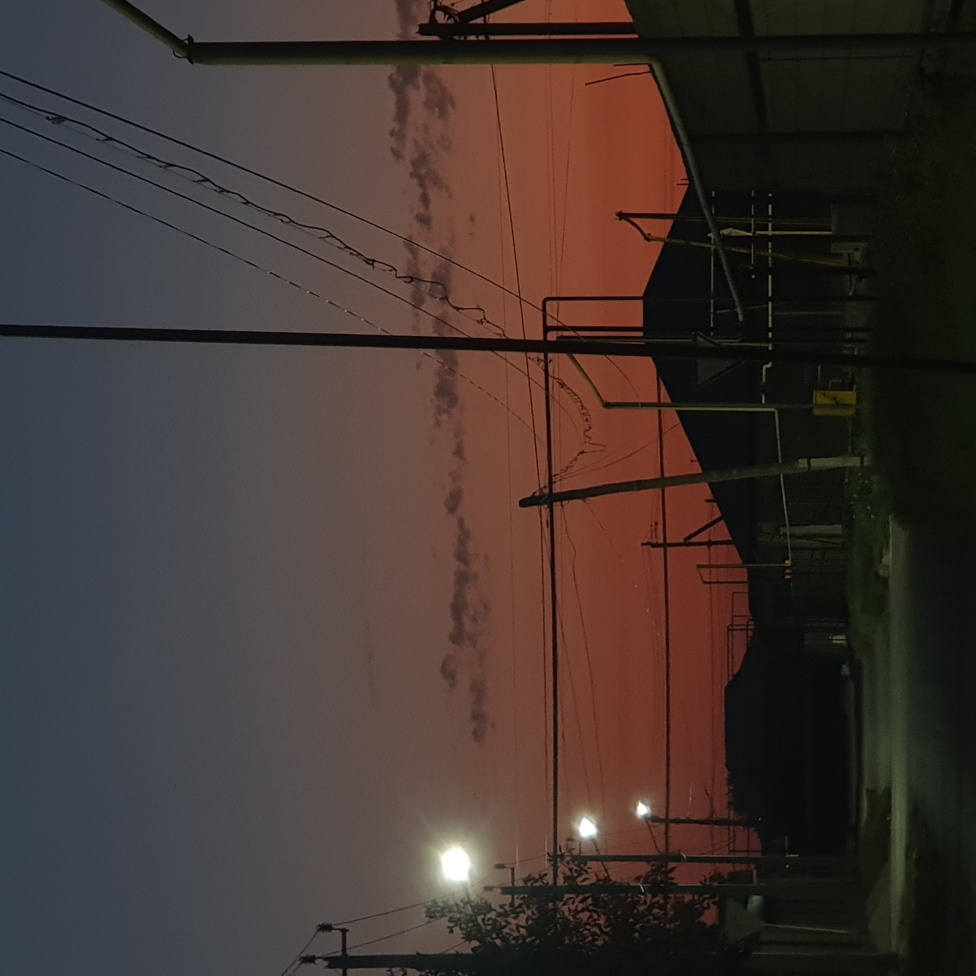The image depicts a street scene at dusk in an unknown location. The sky is a gradient of deep blue to bright orange, with scattered clouds. Several utility poles with wires crisscross the scene. Streetlights are illuminated, casting a soft glow on the road and surrounding structures. On the right side of the image, a building with a dark roof is visible, with some metal piping and a yellow utility box. On the left, a sidewalk and a fence are present. The overall impression is one of a quiet, residential street transitioning from day to night.