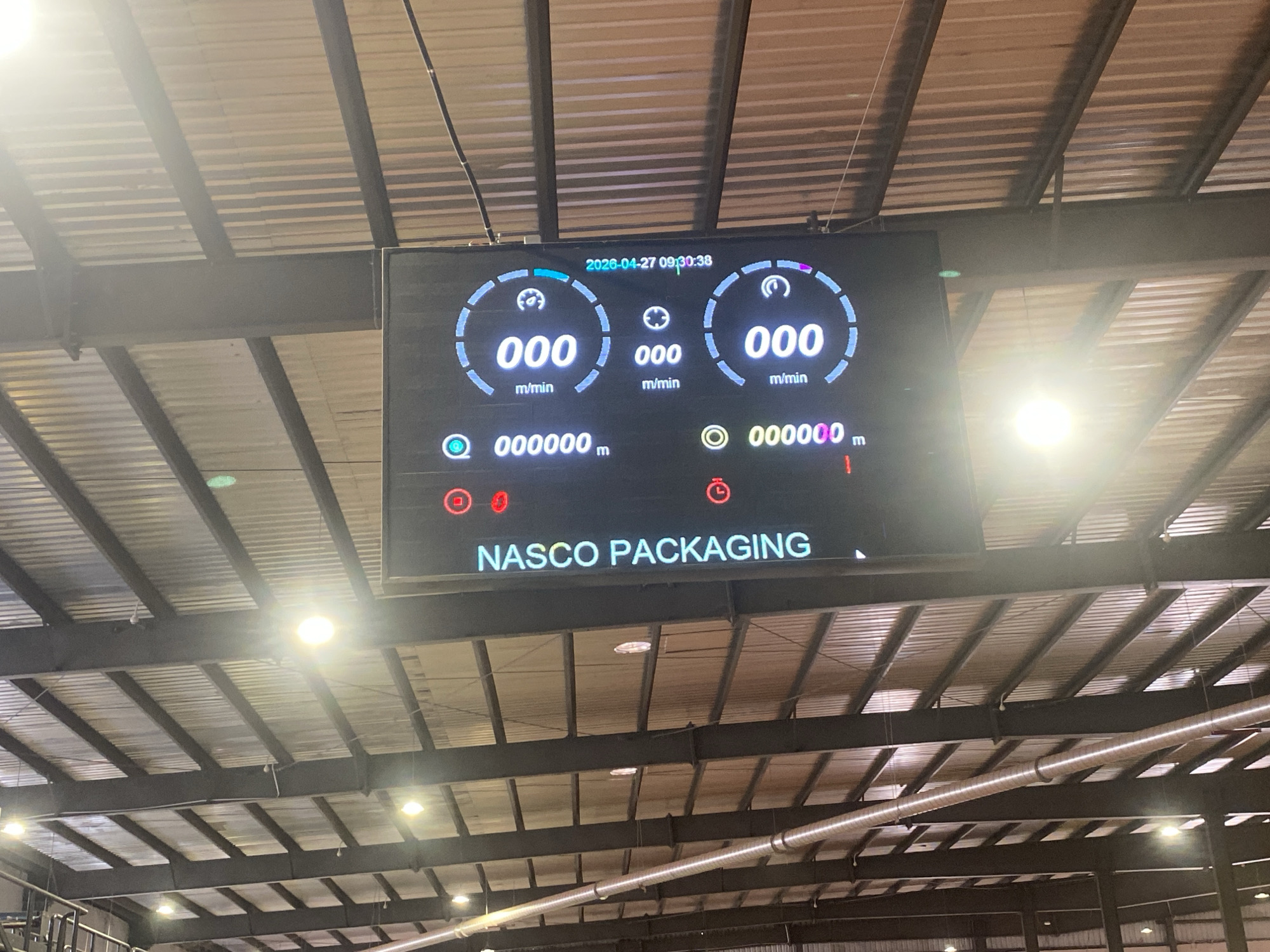 An elevated view captures a digital display screen mounted from the ceiling of an industrial facility in Paipe, Nigeria. The large, rectangular screen features a dark background with bright, digitally rendered information. At the top center, the date and time "2026-04-27 09:30:38" are visible. Below this, three central gauges display "000 m/min," indicating speed measurements, likely meters per minute. Beneath these, two prominent readouts show "000000 m," suggesting total distance or length accumulated. Various smaller icons, including green circles with 'Q' and 'O' symbols, and red circular indicators, are also present, possibly denoting status or alerts. At the bottom of the display, the company name "NASCO PACKAGING" is clearly visible.
The screen is positioned beneath a high industrial ceiling constructed from dark metal I-beams and corrugated metal panels. Several bright, circular industrial light fixtures illuminate the space, with one causing a notable glare on the screen. A silver, cylindrical HVAC duct runs horizontally across the upper background. The overall scene depicts an indoor manufacturing or packaging plant, likely equipped with real-time production monitoring systems, currently showing an idle or reset state.