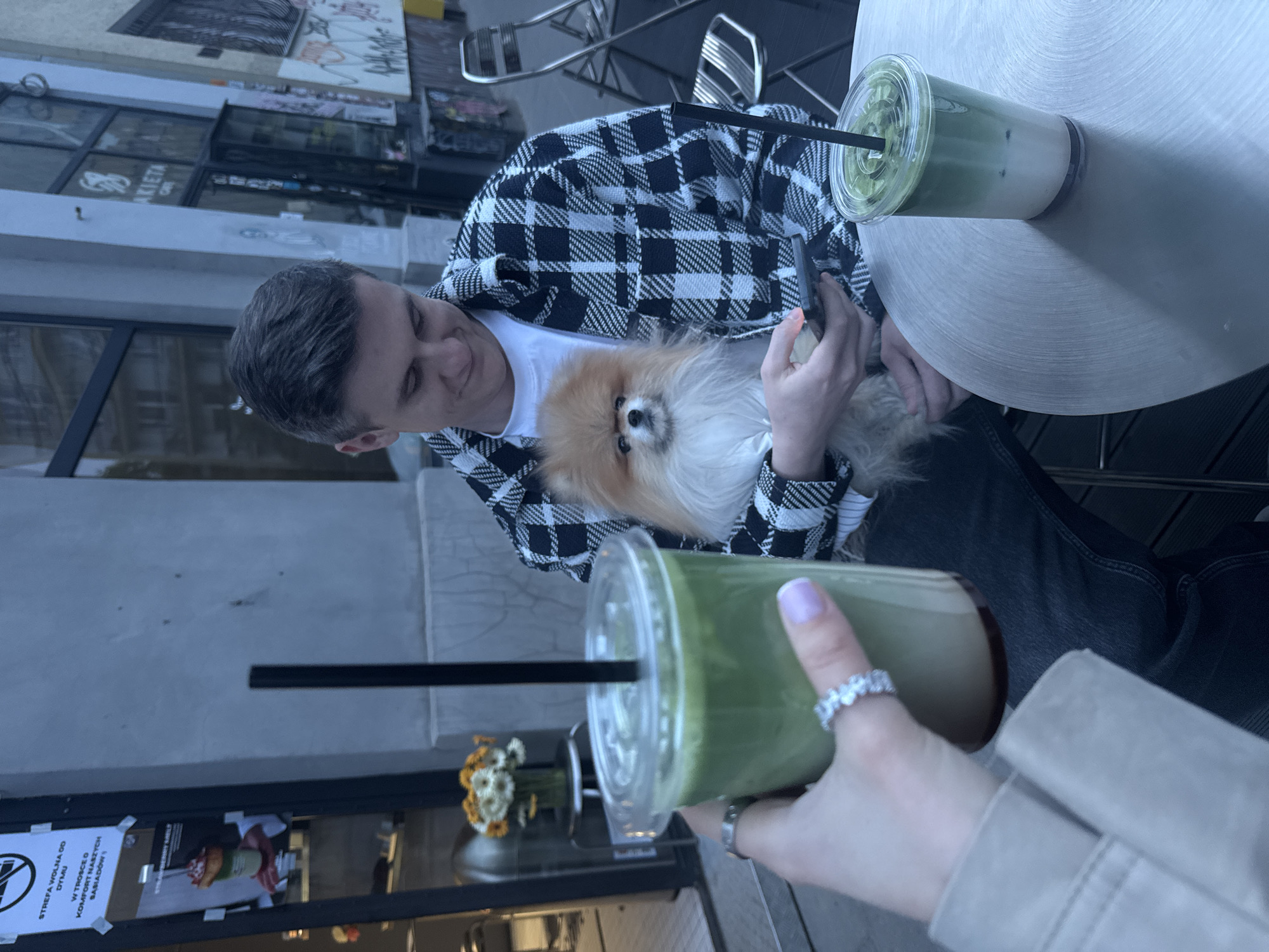 A man wearing a black-and-white plaid jacket and jeans sits at an outdoor table in Warsaw, Poland, holding a small, fluffy orange dog. He is looking down at the dog. There is a cup of matcha latte on the table next to him. In the foreground, a woman's hand with a ring is holding another cup of matcha latte. A storefront with floral decorations is visible in the background, along with a sign in Polish. The table is round and metallic.