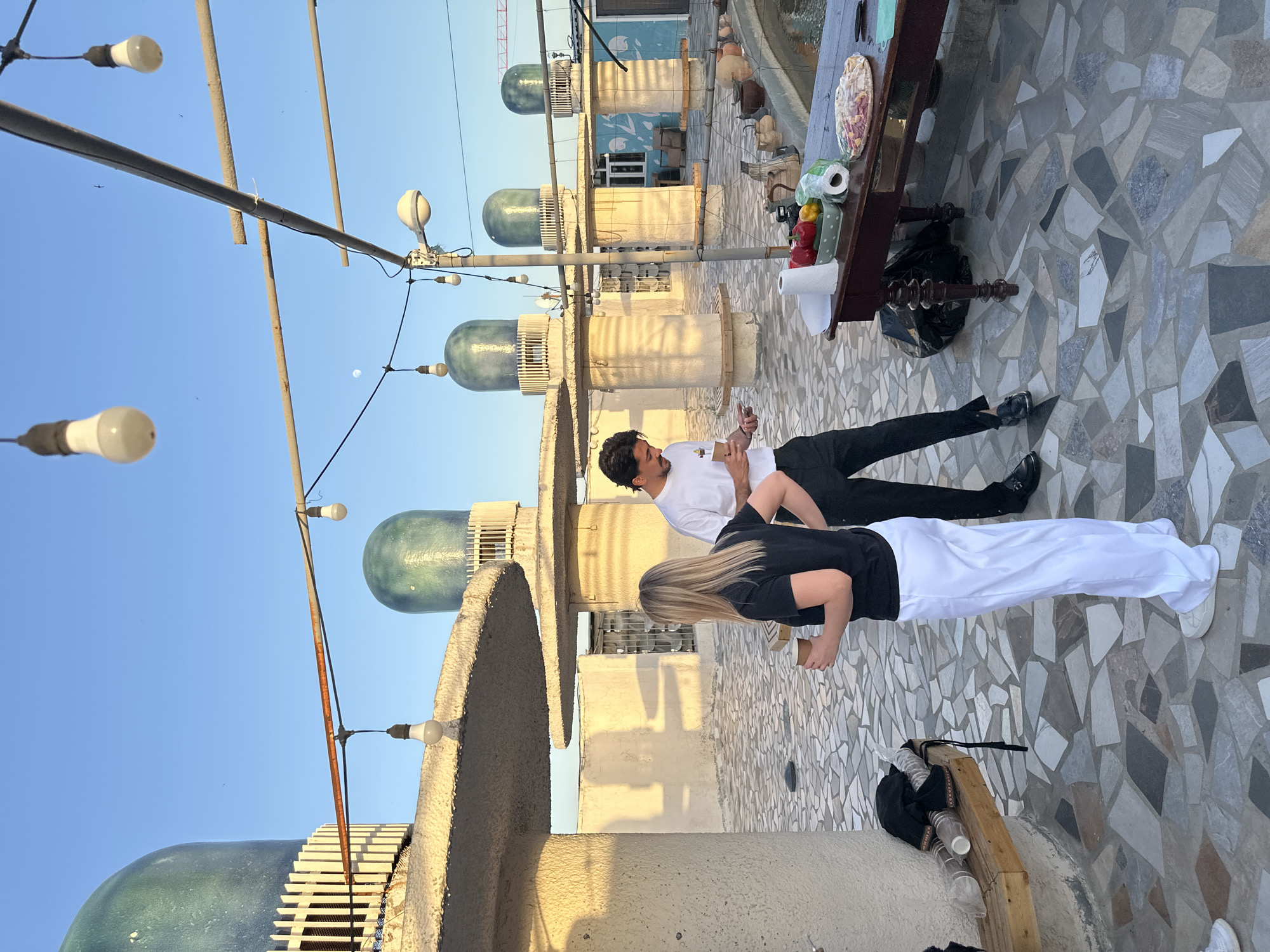 The photograph appears to be taken on a rooftop in Tashkent, Uzbekistan. The rooftop is paved with irregularly shaped stones. Several domed structures are along one side of the rooftop, and string lights are strung above.
Two people are present in the image. One individual is wearing a white T-shirt and black trousers and appears to be looking at a card or small object. The other individual is wearing a black top and white trousers.
A dark wooden table is visible, bearing food and a roll of paper towels. A black plastic bag sits near the table.