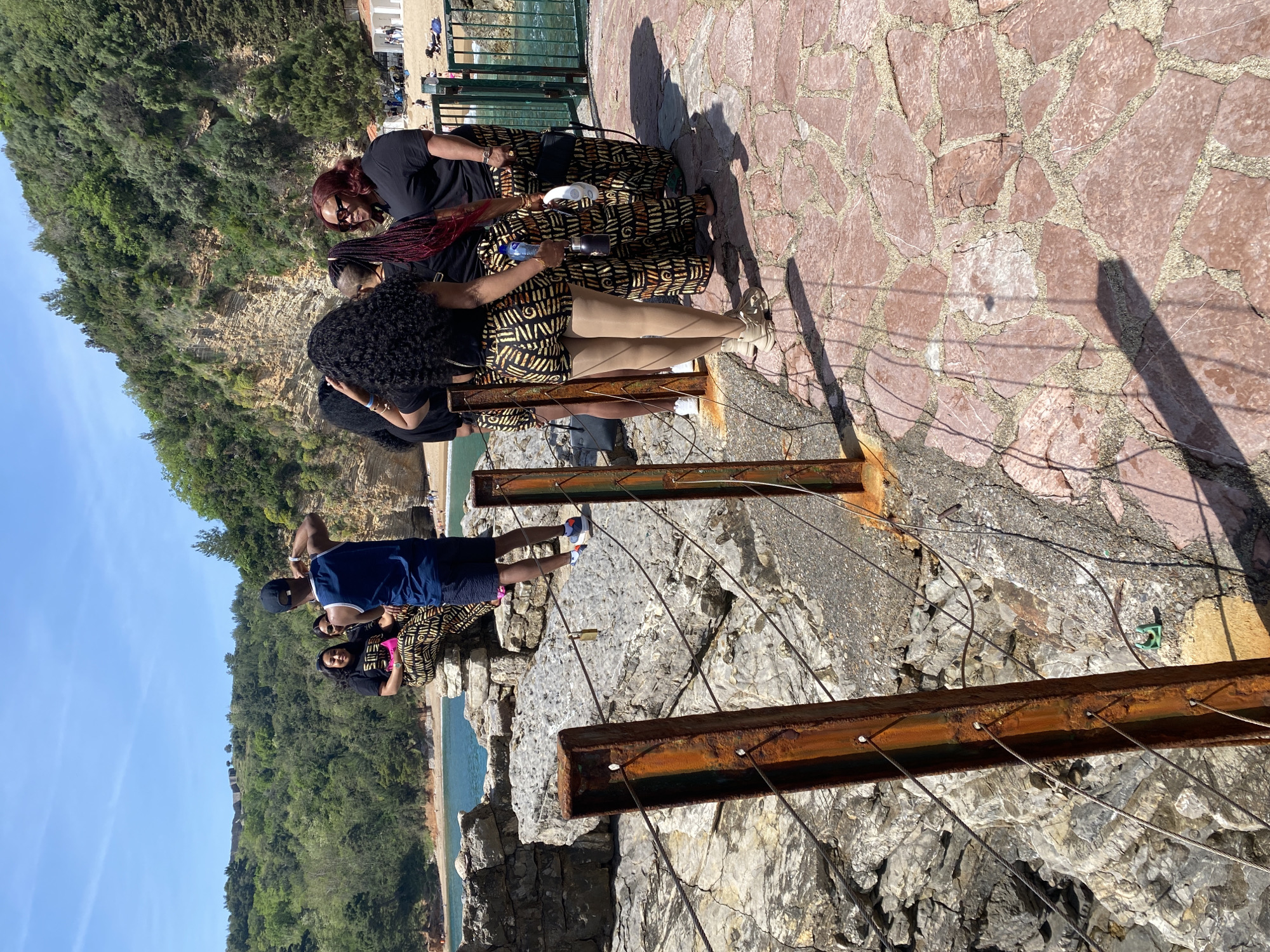 This photograph depicts a group of people on an observation point in the city of Budva, Montenegro. The viewpoint is built on rocky terrain, and a rusted metal structure with supporting cables serves as a safety barrier. In the background, the Adriatic Sea is visible, along with a beach and lush greenery covering cliffs. The sky is clear and blue.
The group of people appear to be tourists or locals. One woman with red braided hair is holding a water bottle. Two people are wearing similar brown-and-black patterned outfits. A man in a navy blue shirt and shorts stands closer to the barrier. A child wearing pink stands behind the man. The viewpoint is constructed of reddish-brown stones.