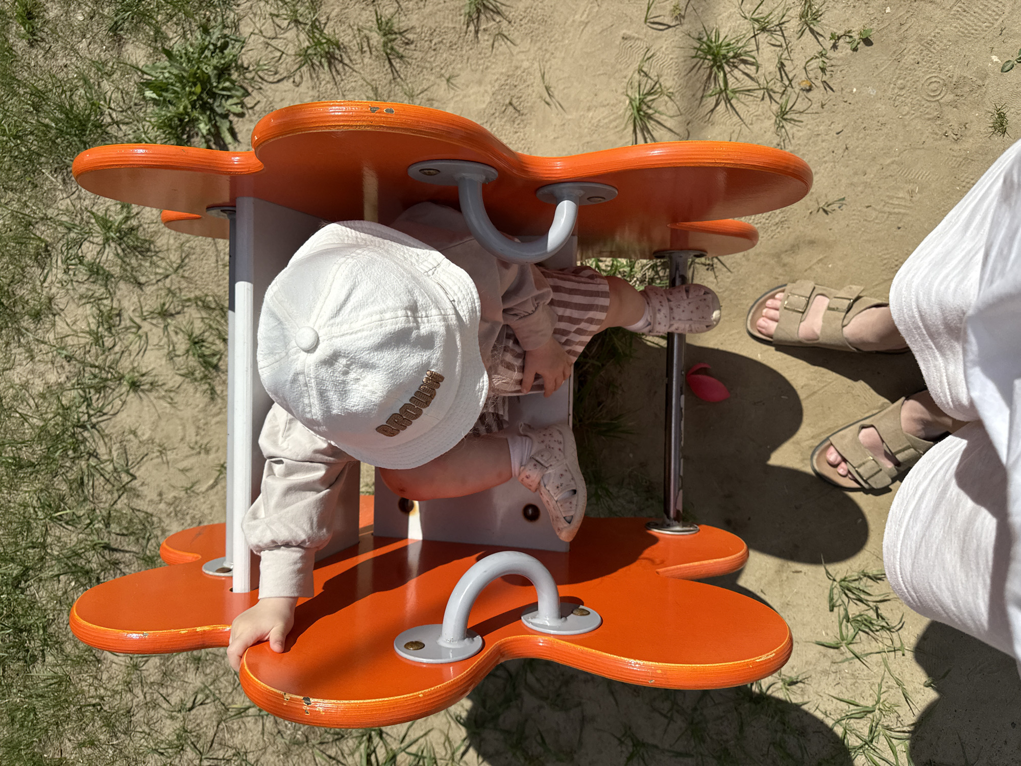 Captured from an overhead perspective, the scene features a child on an orange flower-shaped carousel. The child is wearing a white cap with the word "BROOKLYN" embroidered, along with a beige long-sleeved shirt and striped pants. The child's hand is holding onto the carousel's edge. The carousel is set on a sandy surface with patches of grass. Part of an adult, possibly a parent, wearing sandals and white pants, is visible on the right. The image was taken outdoors under natural lighting. It is unknown if this image was taken in Vladimir, Russia.