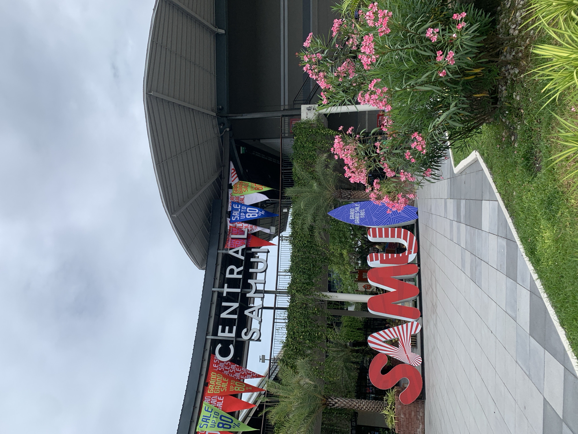 The image shows the entrance to Central Samui, a shopping mall in the Ko Samui District of Thailand. A large sign with the words "CENTRAL SAMUI" is visible, adorned with triangular banners advertising a "GRAND SALE UP TO 80%". In front of the mall, large red letters spell out "SAMUI", with the letter "A" featuring a sunburst pattern. A decorative surfboard with a blue and white zigzag pattern is also present, along with palm trees and a blooming bush with pink flowers. The ground is paved with light-colored tiles and bordered by green grass. The sky is overcast. There are no people visible in the image.