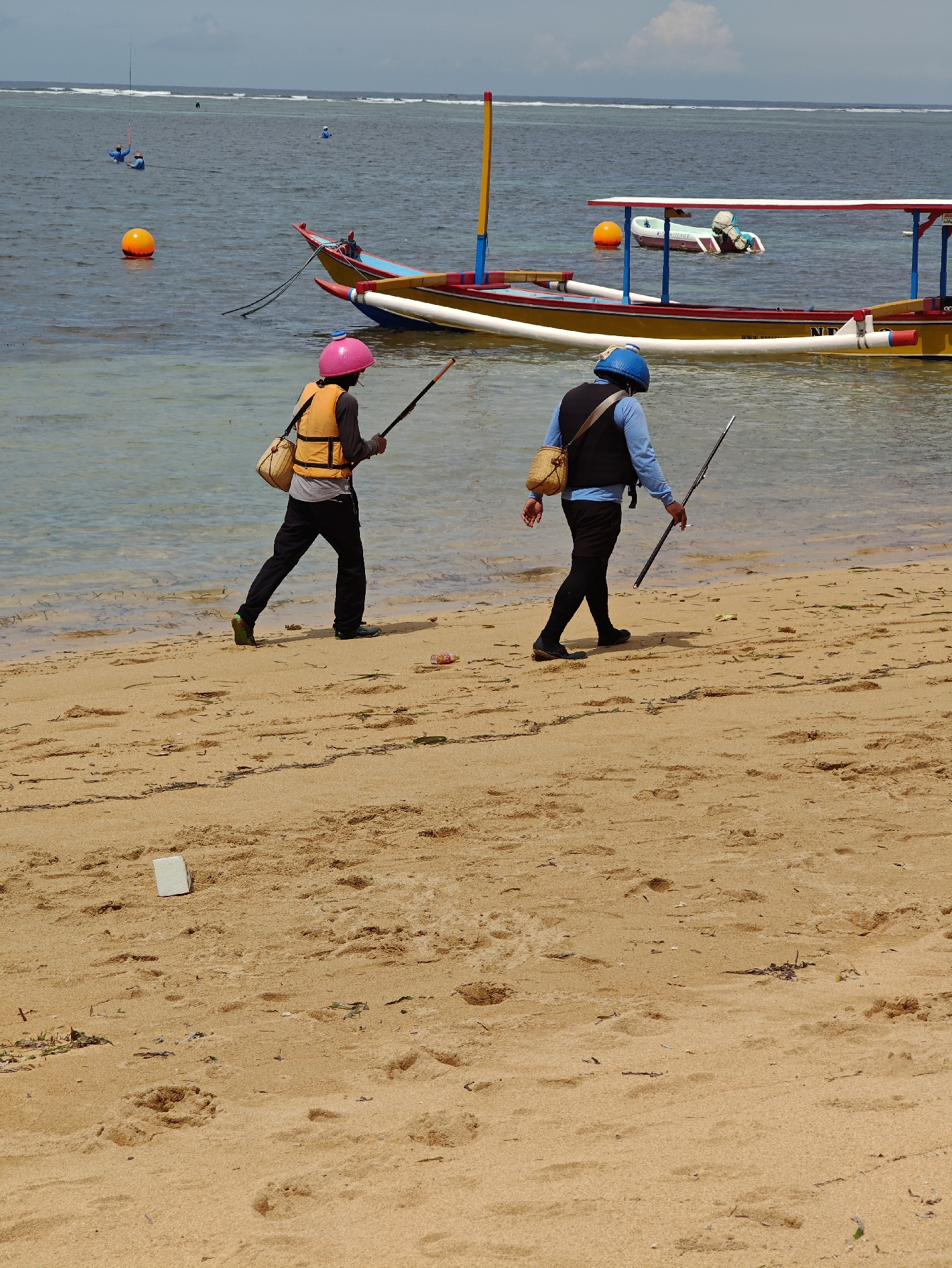 Two individuals, seen from behind, walk along a light brown sandy beach beside the ocean in Denpasar, Indonesia. The person on the left wears a pink helmet, an orange life vest over a grey long-sleeve shirt, and black pants, carrying a woven straw bag slung over their shoulder and a long, slender pole or spear in their right hand. The person on the right wears a blue helmet, a dark vest over a light blue long-sleeve shirt, and dark pants, also carrying a similar woven bag and pole. Both appear to be wearing dark foot coverings.
The calm, clear ocean water shows a gradient of light blue-green near the shore, deepening further out. Several traditional Indonesian outrigger boats, known as jukung, are moored in the water. One prominent boat features a yellow hull with red, blue, and white decorative elements, and a white canopy structure. Partial text "NI**" and possibly "Water sp..." is visible on the side of the yellow boat. Orange buoys float in the water, some associated with the boats. In the distance, white breaking waves indicate a reef or shallow area, and smaller boats or individuals on the water are visible. The sandy beach shows footprints and scattered natural debris like seaweed and small sticks, along with some litter such as a plastic bottle and a white block. The scene is brightly illuminated under a sky with scattered clouds, indicating a clear day.