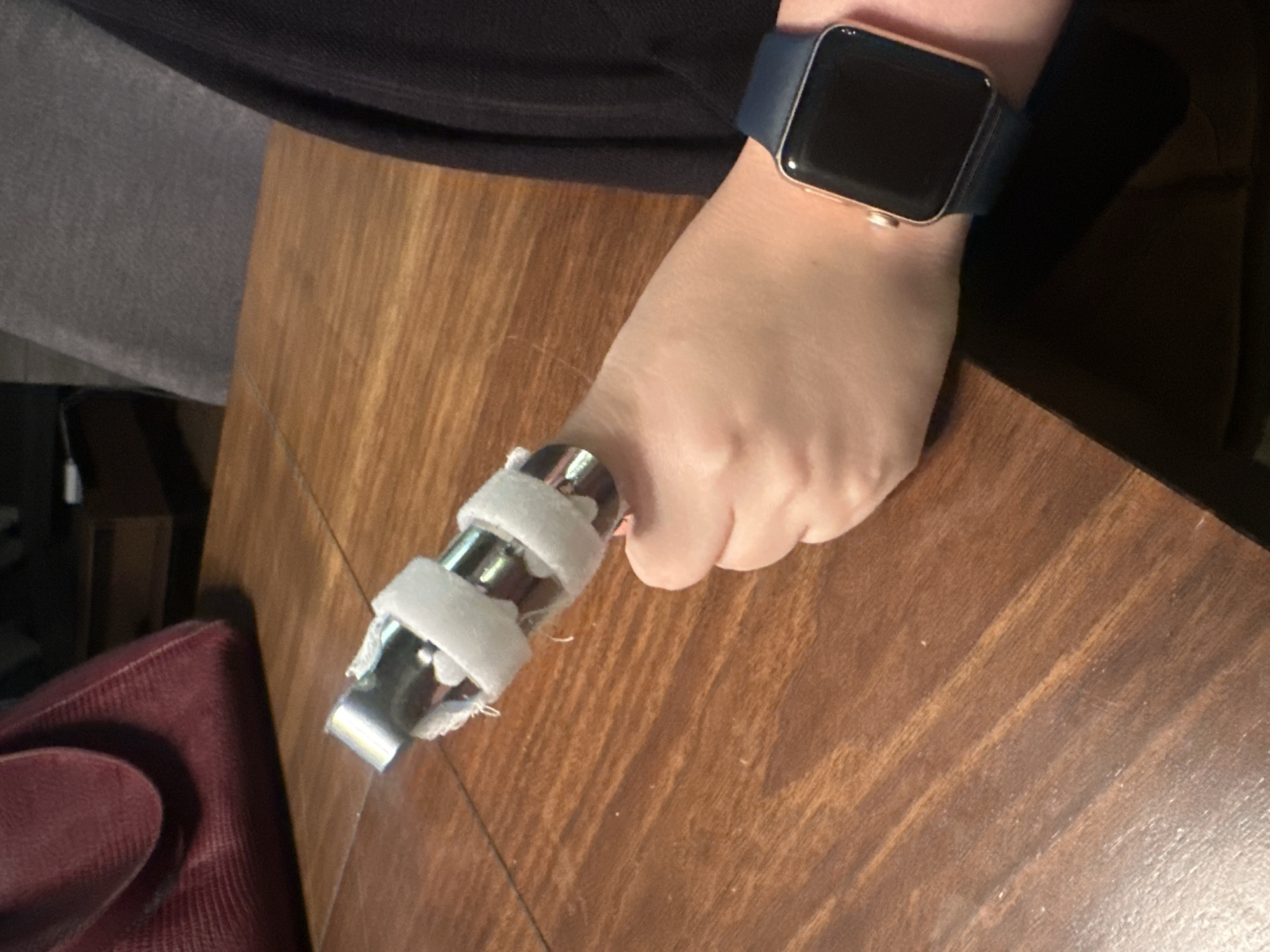 The image shows a close-up of a person's right hand and wrist resting on a wooden table. The index finger is splinted with a metal device wrapped in white material. The person is wearing a dark blue Apple Watch on their left wrist. In the background, a dark red purse and a grey chair are visible. The scene appears to be indoors, possibly at a dining table. There is no indication of the specific location within Huixquilucan de Degollado, Mexico.