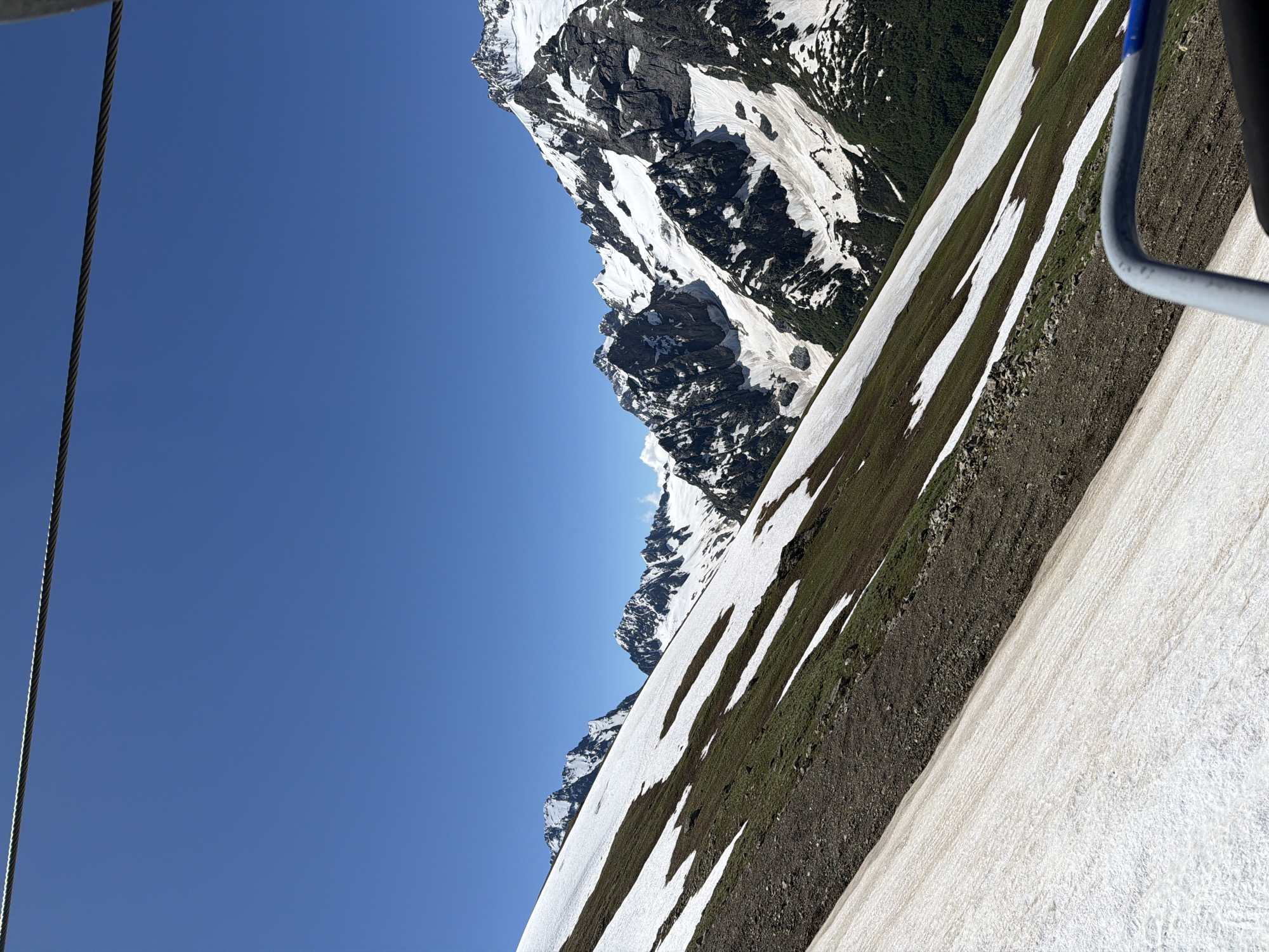 The image depicts a scenic mountain landscape under a clear blue sky. A thick steel cable stretches across the top of the frame, suggesting the presence of a cable car or ski lift. The foreground is dominated by a snow-covered slope, with patches of brown earth and green vegetation visible. This slope is part of a larger mountainside, with more snow-capped peaks in the distance. The mountains are rugged and rocky, with stark contrasts between the white snow and dark rock. A dense green forest is visible in the valley between the mountains. The scene appears to be from a high vantage point, possibly from the cable car itself, looking out over the vast mountainous terrain. The overall impression is one of natural beauty and grandeur, typical of a high-altitude alpine environment. The location is specified as Verkhne-Teberdinskoye Rural Settlement, Russia, suggesting a mountainous region within that administrative area.