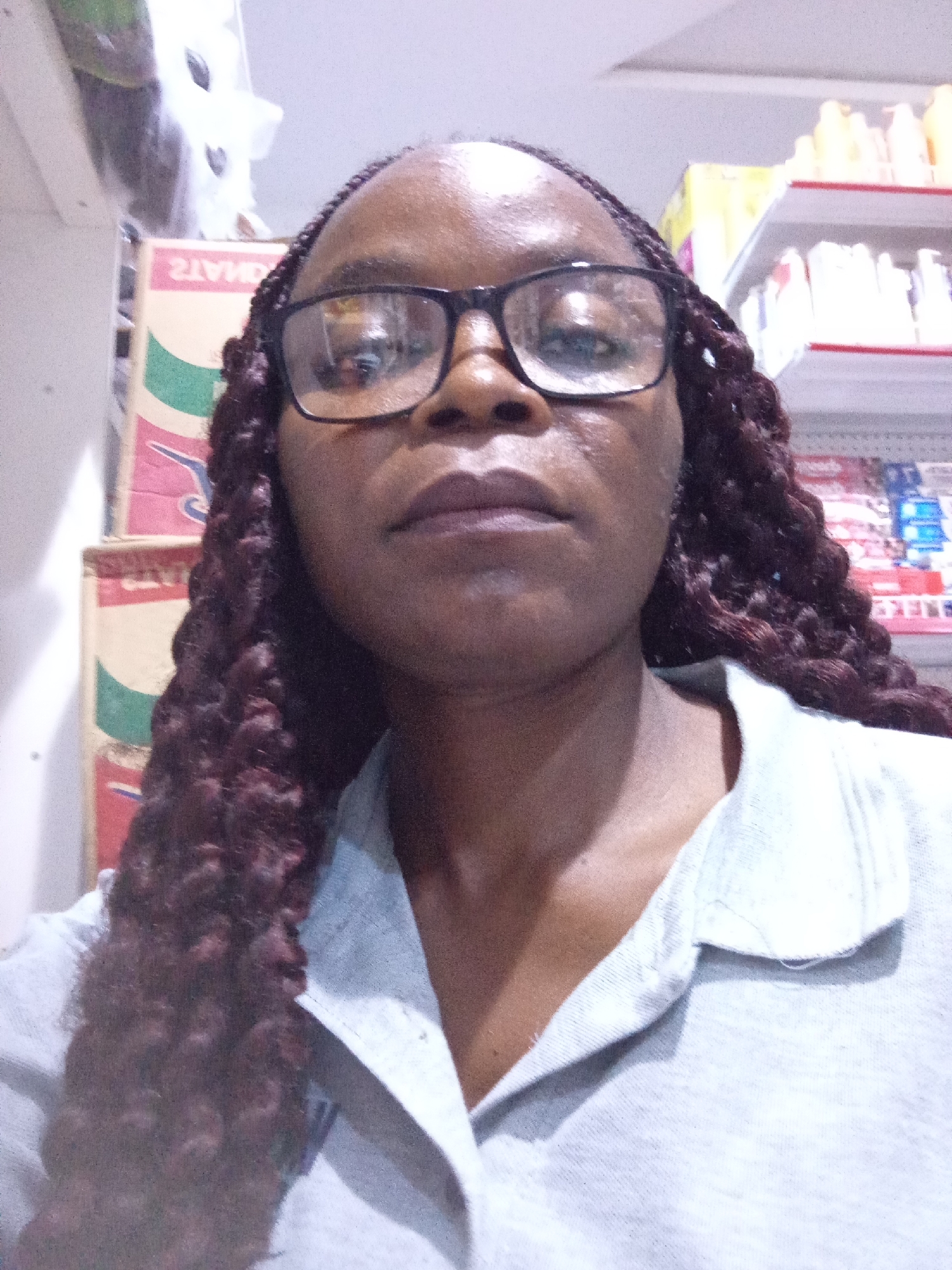 A close-up selfie shows a Black woman wearing black-rimmed glasses and a light blue collared shirt. Her dark, braided hair is visible on the sides of her head. The background suggests an indoor setting, possibly a store, with shelves of products faintly visible. Behind her left shoulder, stacks of boxes with colorful labels, including one with "21MD" printed in reverse, are discernible. The lighting appears to be artificial, indicating it could be daytime or nighttime. No specific emotions are strongly conveyed, but her expression is neutral. The location context is Howayi, Nigeria.