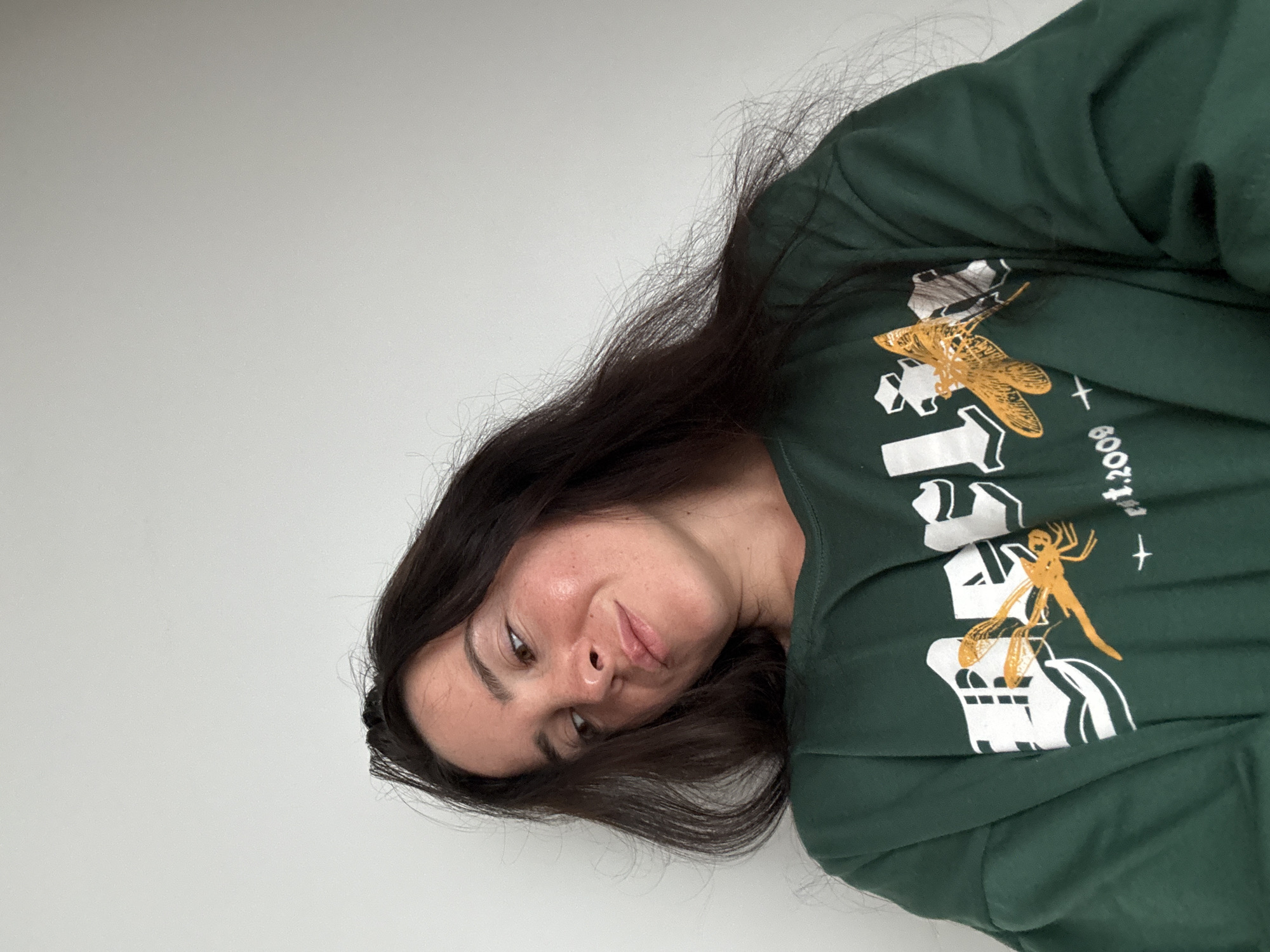 A young woman with long, dark brown hair is shown from the chest up, looking down and to her left. She is wearing a dark green sweatshirt with a graphic design featuring white lettering and two orange dragonflies. The background is a plain, light-colored wall. The scene appears to be indoors, with soft lighting. The woman's facial features are visible, and she has a light complexion with some freckles. The image is a portrait-style shot, with the camera positioned slightly above and to the side of the subject.