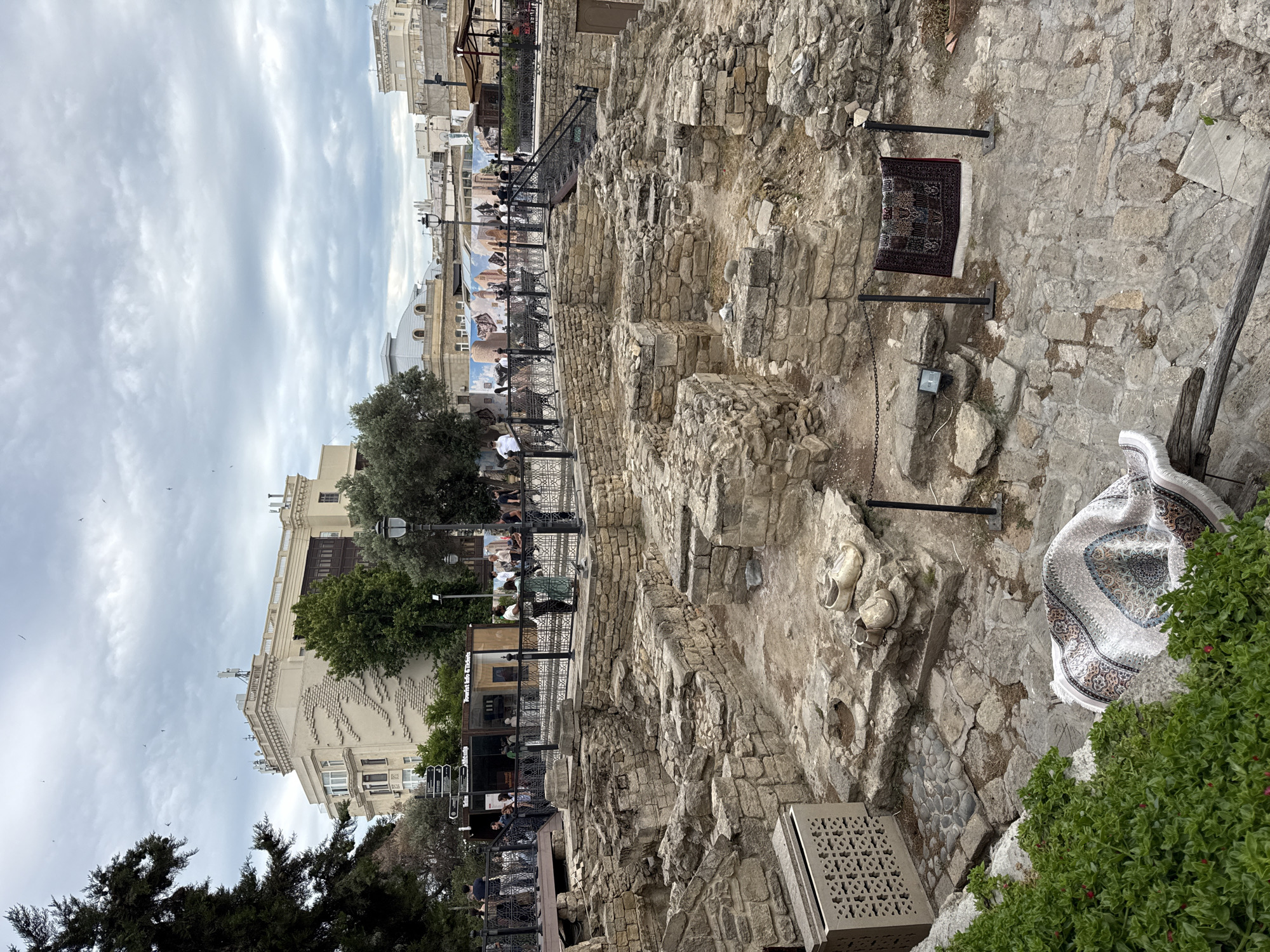 The image depicts the archaeological site of the Shirvanshahs' Palace in the Old City of Baku, Sabail Raion, Azerbaijan. The foreground features the stone ruins of ancient structures, with exposed foundations and walls made of rough-hewn stones. Several architectural elements, such as arched doorways and remnants of columns, are visible. In the middle ground, a decorative rug is displayed, hung between two posts. Nearby, a larger rug lies on the ground, partially unfurled. Two pairs of worn, light-colored shoes are placed on a stone ledge. A decorative stone planter with intricate carvings is situated to the left.
In the background, a bustling scene unfolds. A raised walkway with a black metal railing is lined with people observing the ruins. Beyond the walkway, a modern glass-fronted building with signage that includes "Tourist Info & Tickets" is visible, suggesting a visitor center. Further in the background, several multi-story buildings with distinct architectural styles can be seen, including a large, pale building with rows of windows and decorative elements, and other structures with varying rooflines and facade designs. The sky is overcast with scattered clouds, and a few birds are visible flying. The overall impression is a juxtaposition of ancient history and modern urban life.