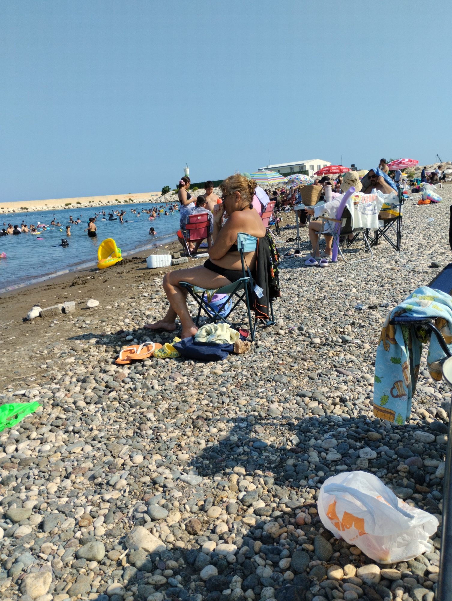 The image depicts a crowded beach in Erdemli, Turkey, under a clear blue sky. The foreground is dominated by a pebble-strewn beach with several people relaxing in beach chairs. A woman in a black swimsuit sits in a folding chair, facing away from the camera. In front of her, on the pebbles, are a pair of orange flip-flops and yellow water shoes. To the right, a white plastic bag with orange markings is visible. Further back, more people are seated on the beach, many under colorful umbrellas. A body of water, likely the sea, is populated with numerous swimmers. Along the water's edge, a yellow inflatable toy is present. In the distance, a breakwater extends into the sea, with a white lighthouse visible on it, and some buildings are situated beyond the beach. The overall scene conveys a typical summer day at a popular coastal location.
