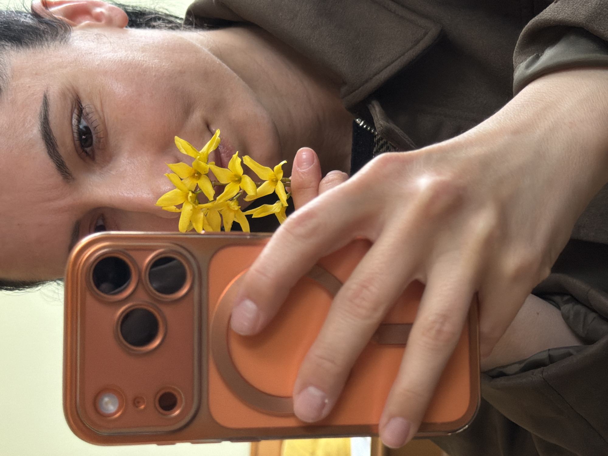 A woman is taking a selfie with her phone, holding a small cluster of yellow flowers near her face. The phone has a distinctive orange case with a circular grip on the back. She is wearing a brown jacket. The background is a light cream color. The photo does not contain enough information to identify the location as Pleven, Bulgaria.