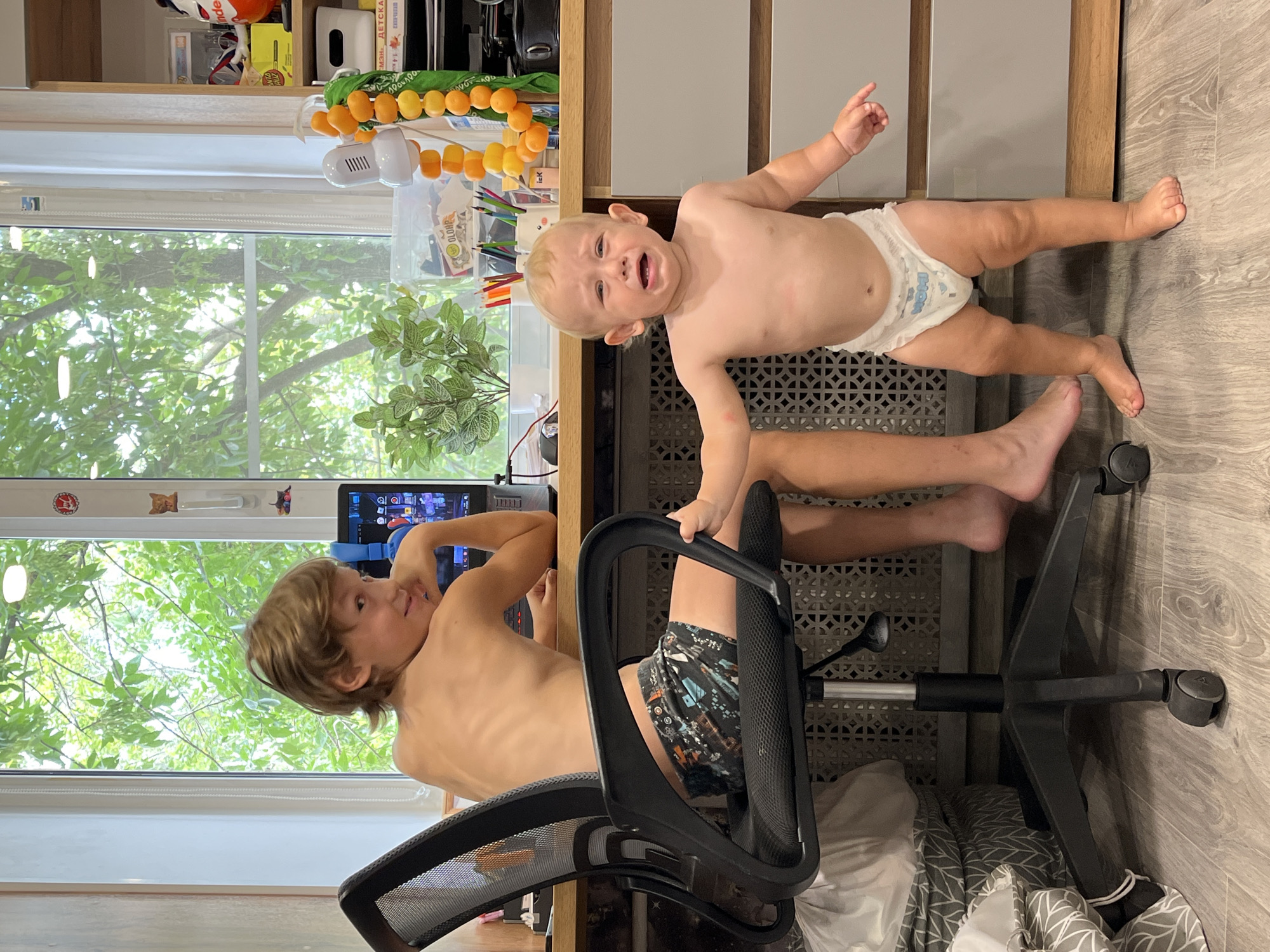 A young child, possibly a toddler, is crying while being held up by an older child. The older child is shirtless and wearing patterned shorts, and appears to be sitting in a black office chair. The scene takes place indoors, with a desk, shelves, and a window visible. The window shows trees and greenery, suggesting it is daytime and possibly a warm season. On the shelves are various objects, including what appear to be toys and art supplies. A tablet is on the desk, displaying a colorful screen. The floor is wooden. The overall mood seems to be one of playful chaos or a moment of sibling interaction. Visible text includes what appears to be Russian text on some of the items on the shelf and on the tablet.