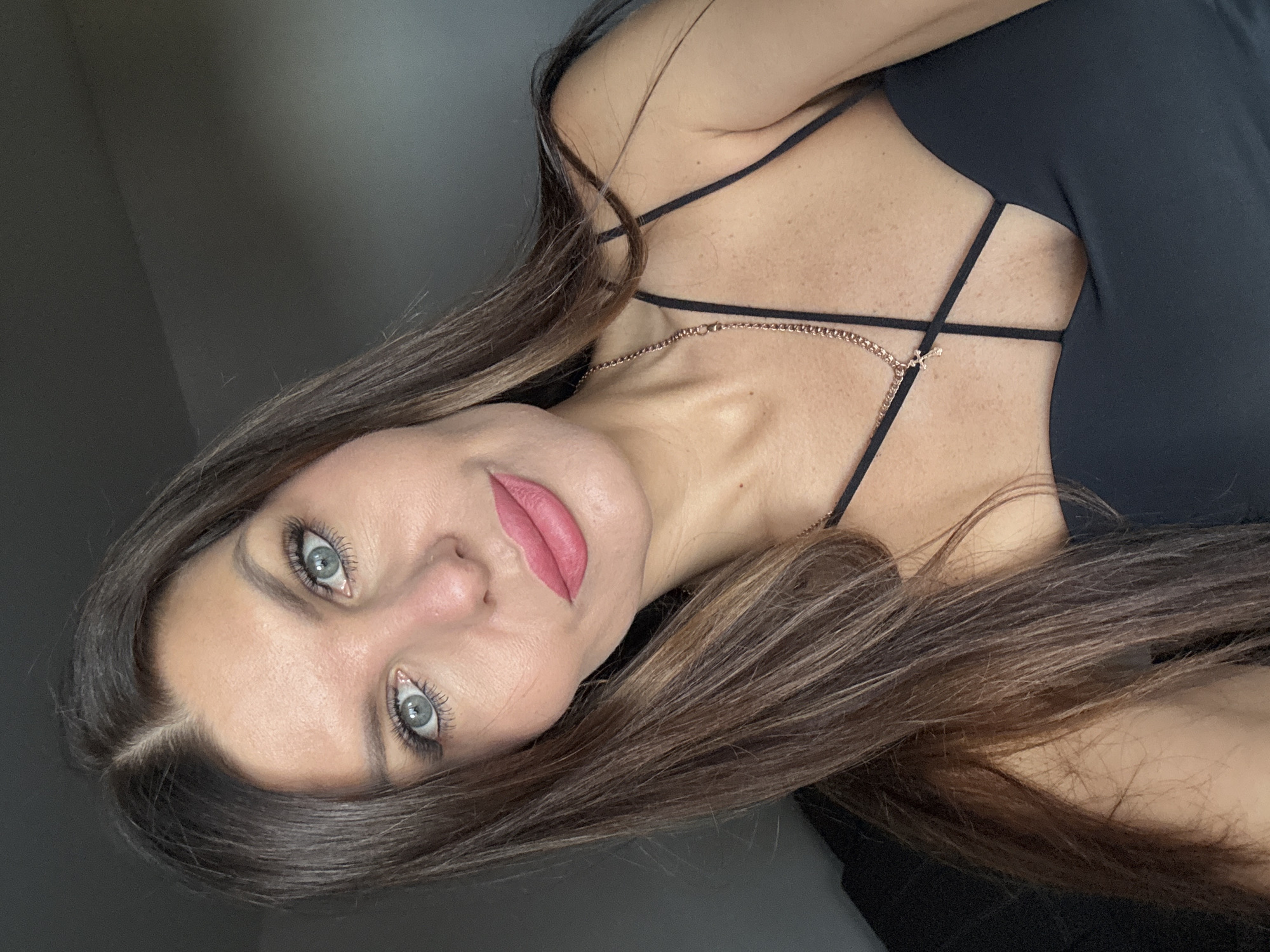 A selfie of a woman with long, dark brown hair and blue eyes. She is wearing a black dress with thin straps and a rose gold necklace with a cross pendant. Her makeup includes pink lipstick and false eyelashes. The background is a plain, dark gray wall. No specific location in Chaikovsky, Russia, can be determined from the image.