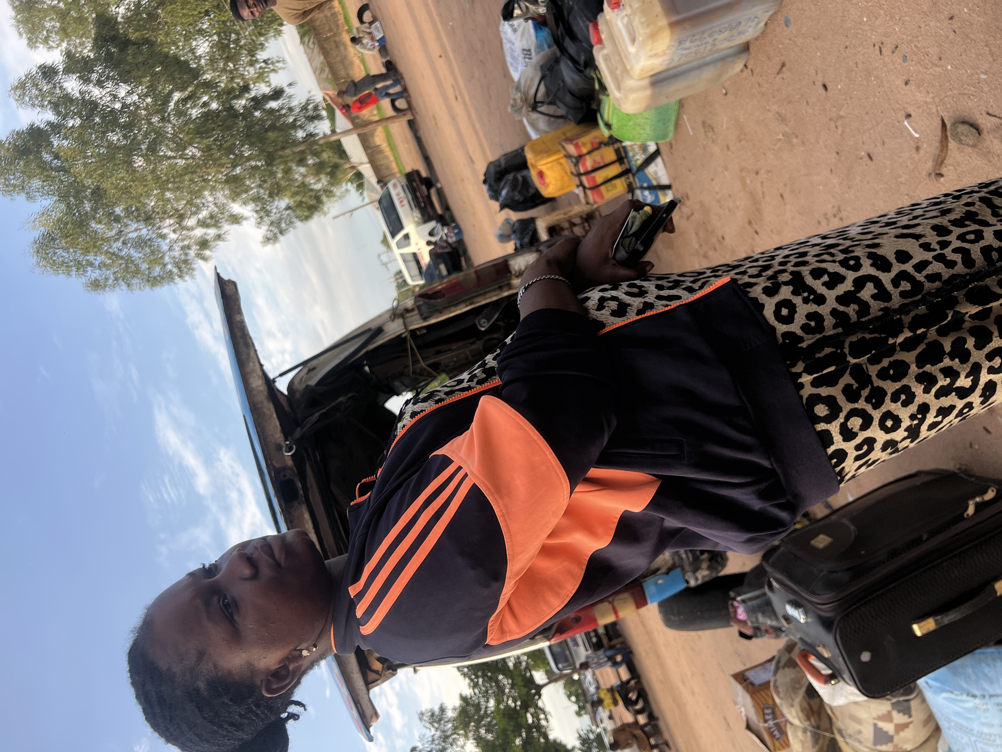 In Gbaza, Nigeria, an outdoor scene unfolds under a bright sky with scattered white clouds. The foreground is dominated by a dark-skinned woman, positioned at a slight angle facing right, with her head turned to look into the mid-distance. She wears a dark blue jacket featuring bright orange stripes on the upper sleeves and orange paneling on the lower arms, over a leopard-print garment. A silver-colored bracelet adorns her right wrist, and she holds a dark-colored smartphone or similar device in her right hand. Small gold-colored hoop earrings are visible on her ears.
The ground is unpaved, consisting of reddish-brown earth with minimal debris. To the woman's left, a black rolling suitcase with a brown handle rests on the ground, next to a large patterned fabric bag and a light blue item. To her right, several translucent plastic jerrycans, filled with a brownish liquid, are stacked on the ground.
In the mid-ground, a white commercial van, marked with "TYTC" or similar text on its side, is parked. Luggage, including numerous black plastic bags and yellow containers, is piled on the ground near the van. Behind the foreground woman, the open rear hatch of a vehicle with a blue roof/trim is visible.
Several individuals are present in the mid-to-background. On the left, a person in a light brown garment sits on a low object. Further right, another person in a yellow shirt also sits. In the background, a person in a red shirt stands near other figures and what appears to be a concrete wall. Tall, leafy green trees are prominent on both the left and right sides of the scene, partially obscuring simple, light-colored buildings with corrugated roofs. On the far right, a person in a light brown shirt is partially visible.