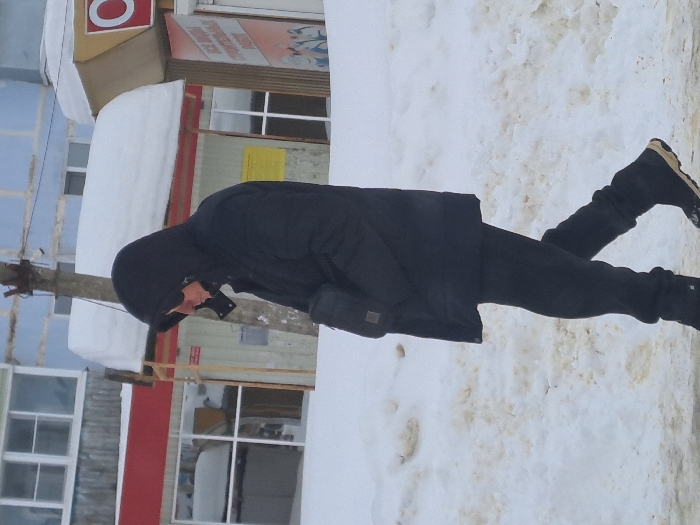 An individual is depicted walking from right to left on a snow-covered street in Sasovo, Russia. The person wears a black hooded puffer jacket with the hood up, dark sunglasses, black gloves, black pants, and black winter boots with a tan sole. A black shoulder bag is visible on their right hip. The ground is covered in white and discolored snow, indicating a winter environment. In the background, multiple buildings are visible, including one with a light blue upper facade, a wooden lower section, and windows, along with a red awning piled with snow. Adjacent to this, another commercial building or shop entrance features a yellow awning also covered in snow, and a red sign with a white circular logo (possibly an 'O'). A weathered utility pole stands between the individual and the background structures.