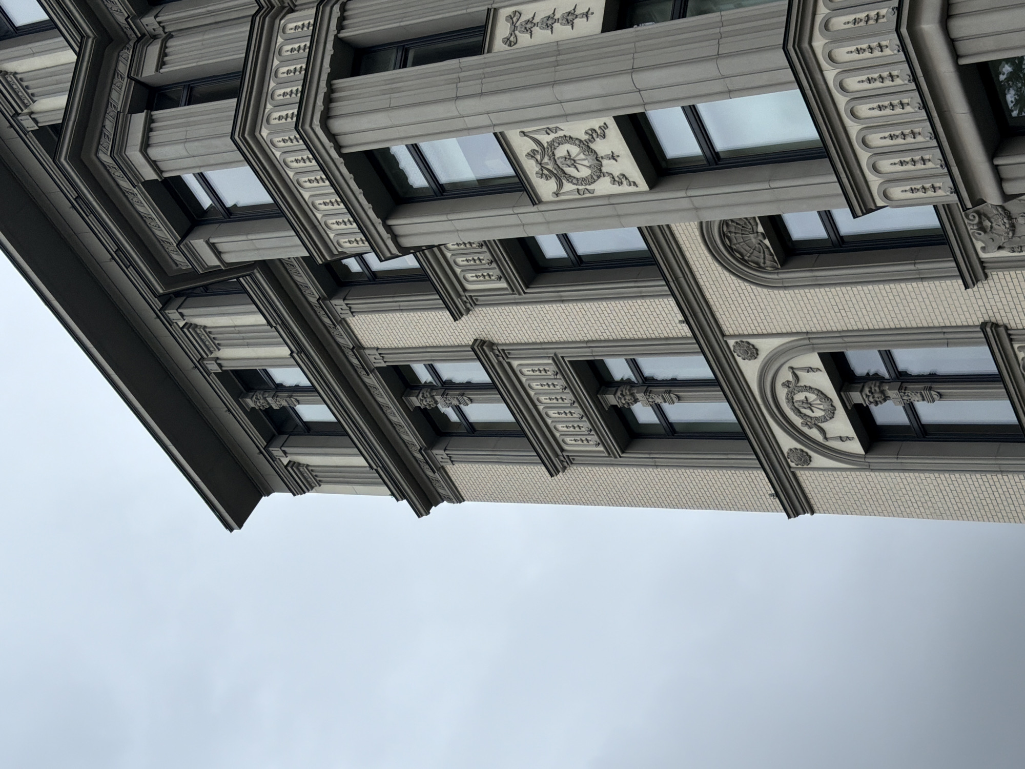 A close-up, upward-angle shot features the ornate facade of a light-colored brick building with grey stone architectural embellishments against a cloudy sky. The building exhibits classical architectural elements such as pilasters, decorative reliefs, and recessed window frames with dark panes. The composition emphasizes the detailed stonework and the contrast between the building's facade and the muted sky. The scene is devoid of people or any discernable activity. The style suggests a historic European city, possibly from the late 19th or early 20th century.