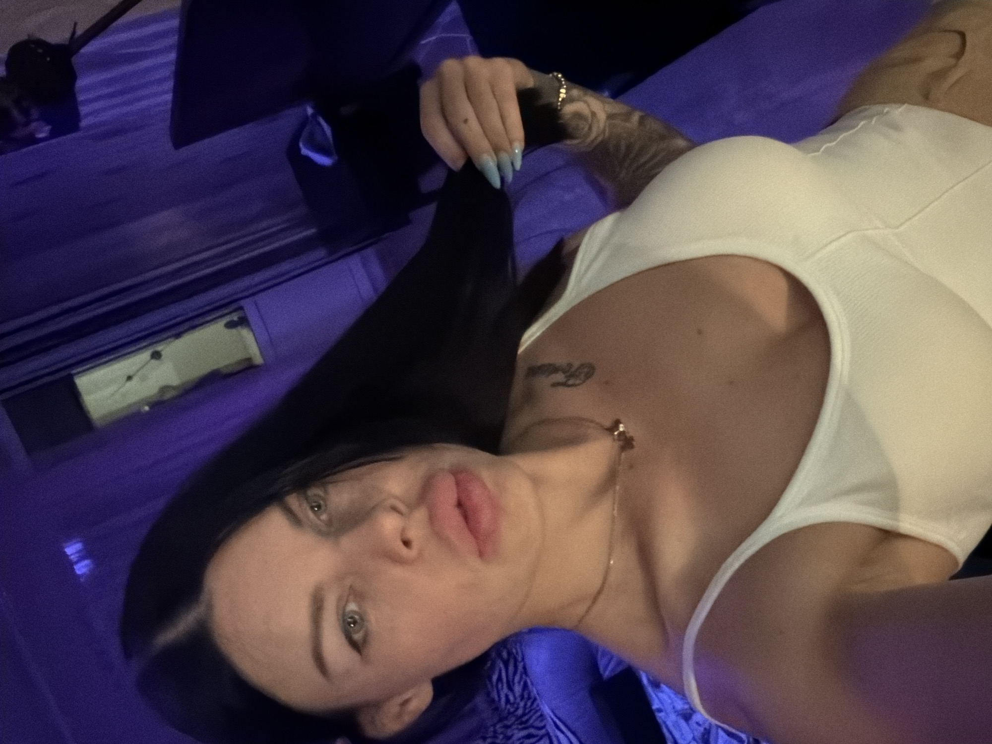 A young woman with long dark hair and striking features is captured in a close-up, selfie-style photograph. She is wearing a white tank top and a delicate necklace. Her expression is direct and engaging, with a hint of a pout. The background is dominated by deep purple hues, suggesting a dimly lit, intimate setting. The lighting casts a subtle glow on her skin and hair, creating a dramatic and alluring atmosphere. The overall impression is one of confidence and allure, with the focus solely on the woman and her captivating presence. The image was taken in an indoor environment, likely a private space, and does not provide any discernible clues about its specific location within the city of Ufa, Russia.
