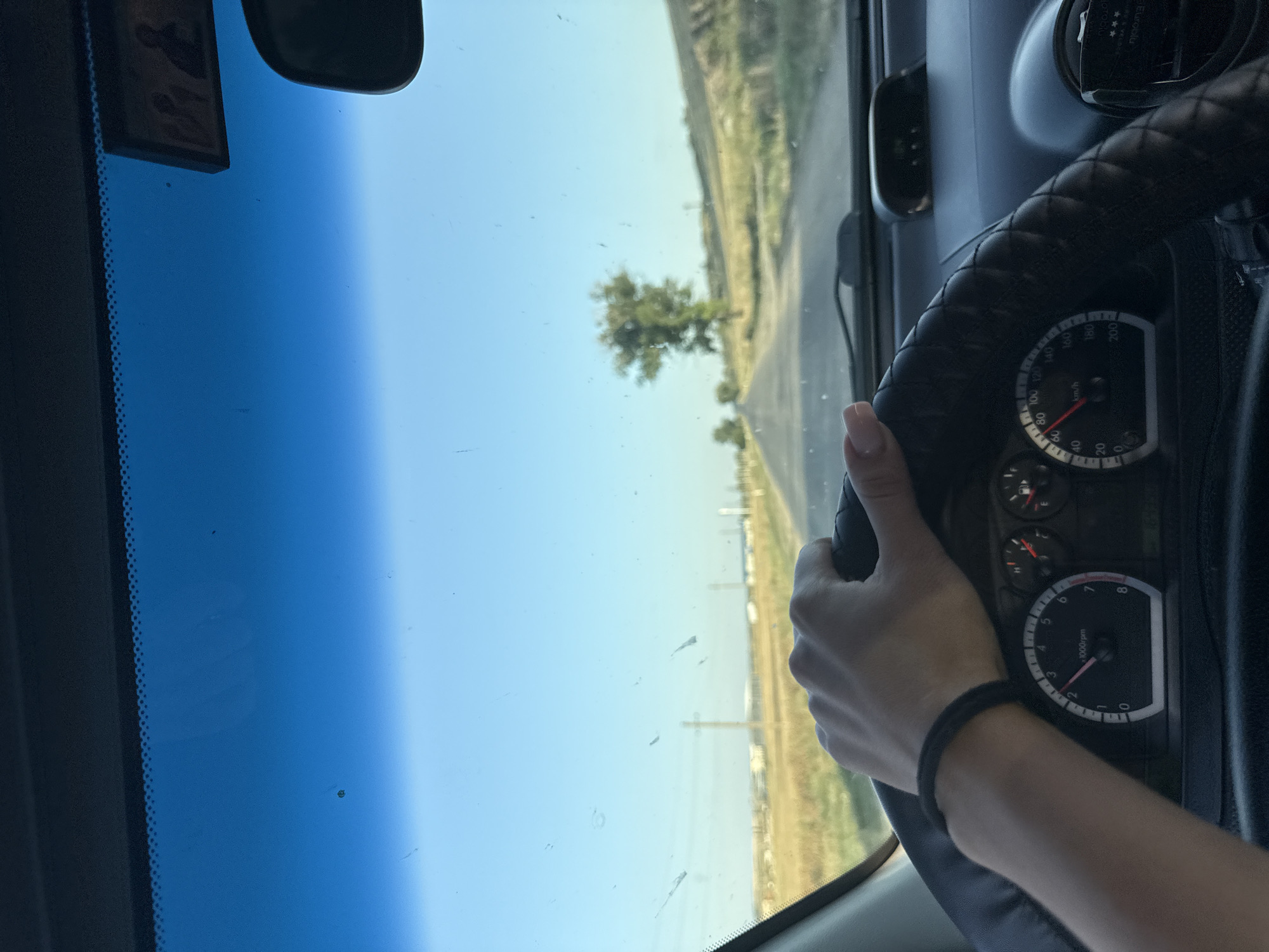 The image, captured from inside a vehicle, shows a person's hand gripping the steering wheel. The dashboard, including a tachometer and speedometer, is visible. Outside the car, a road stretches into the distance, flanked by fields and a solitary tree. The sky is clear and blue. The scene appears to be a rural setting. Based on the context of Cimișlia, Moldova, this image likely depicts a drive on a road outside the city.