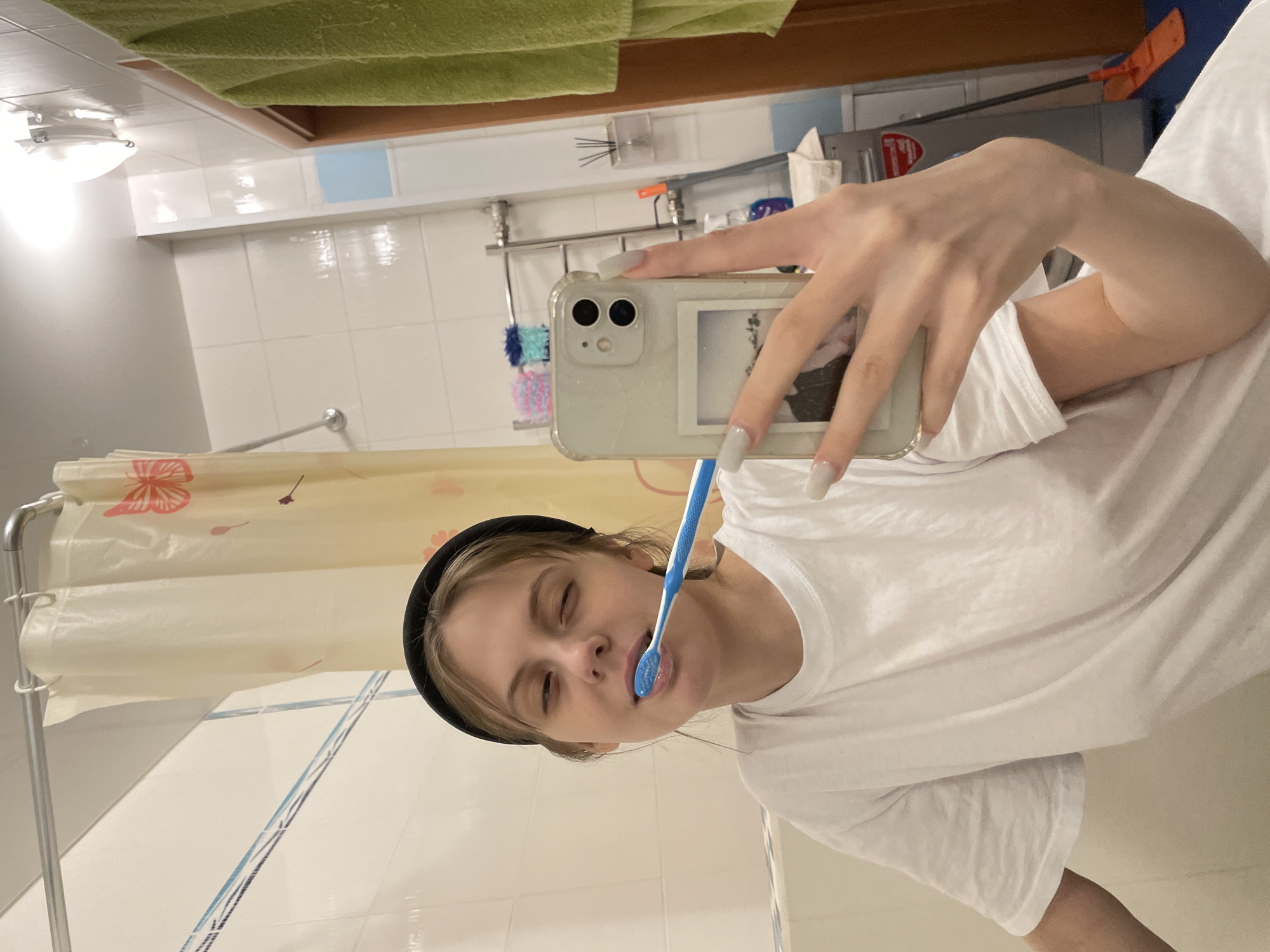 A woman is taking a selfie while brushing her teeth in a bathroom. She is wearing a white t-shirt and a black headband. The bathroom has white tile walls, a shower curtain with a floral design, and a green towel hanging above. There are toiletries on a shelf in the background. The location is likely a private residence in Ufa, Russia.