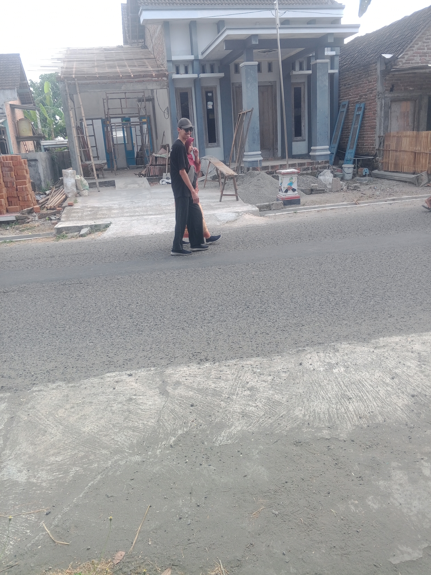 In Lembah, Indonesia, two individuals are walking on a paved road. The person in front is wearing a black t-shirt, black pants, a black cap, and sunglasses. They are walking with their left arm extended slightly forward. Behind them, a person in a pink headscarf and a patterned skirt is walking. Construction is visible on the right side of the image, with a house under renovation. Building materials such as bricks, sand, and scaffolding are present. The environment appears to be a residential area undergoing development.