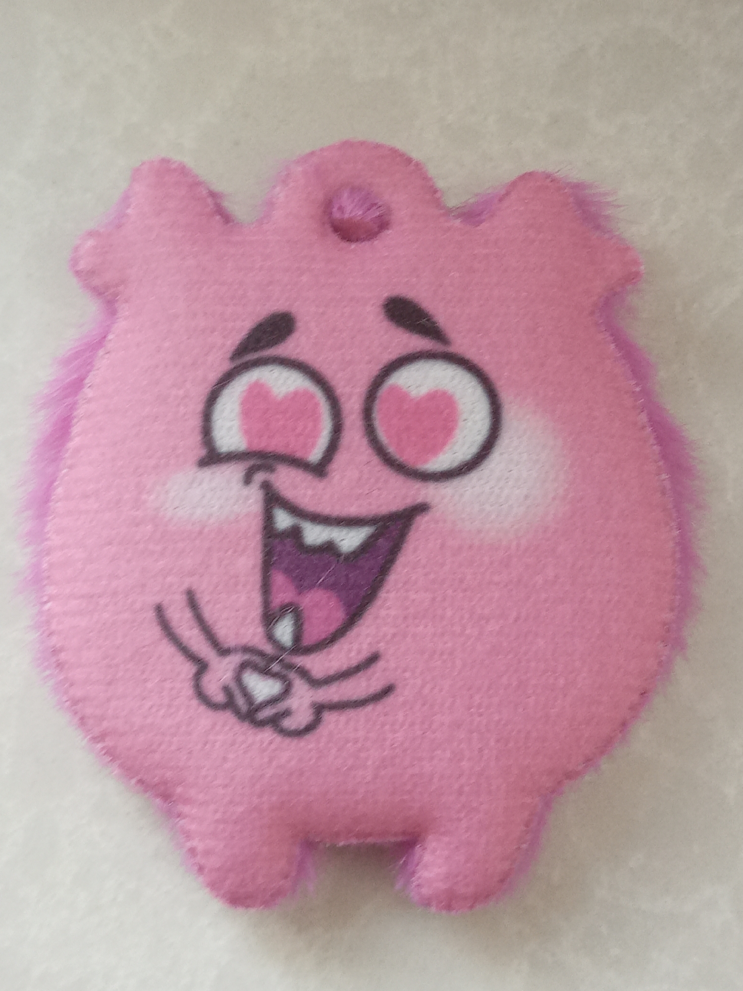 A pink, plush toy resembling a cartoon character with a playful, loving expression is displayed. It has large, heart-shaped eyes, a wide, smiling mouth with visible teeth, and small hands seemingly clasped together. The toy has a fuzzy border around the edges and an opening at the top, possibly for hanging. The object rests on a plain, light-colored surface. There is no indication this image was taken in the city of Elektrostal, Russia.