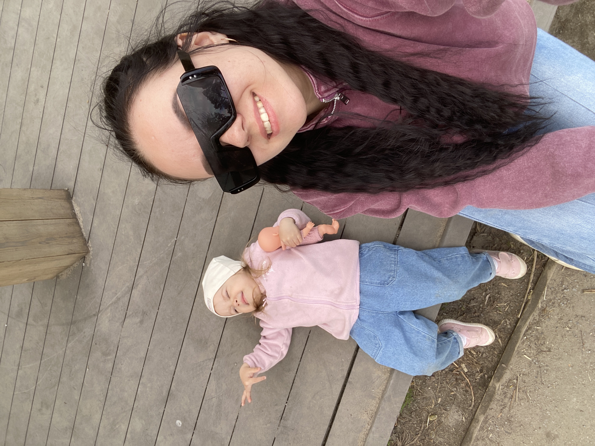 A woman and child are lying on a gray wooden deck in Perm, Russia. The woman, facing the camera, wears a mauve-colored hoodie, blue jeans, and large black sunglasses. She has long, dark, curly hair and is smiling. Beside her, a small child lies on their back, wearing a pink jacket, blue pants, pink shoes, and a white cap. The child holds a doll in their right hand and has their left arm outstretched. The deck appears to be part of an outdoor area, with some dirt or vegetation visible near the edge.