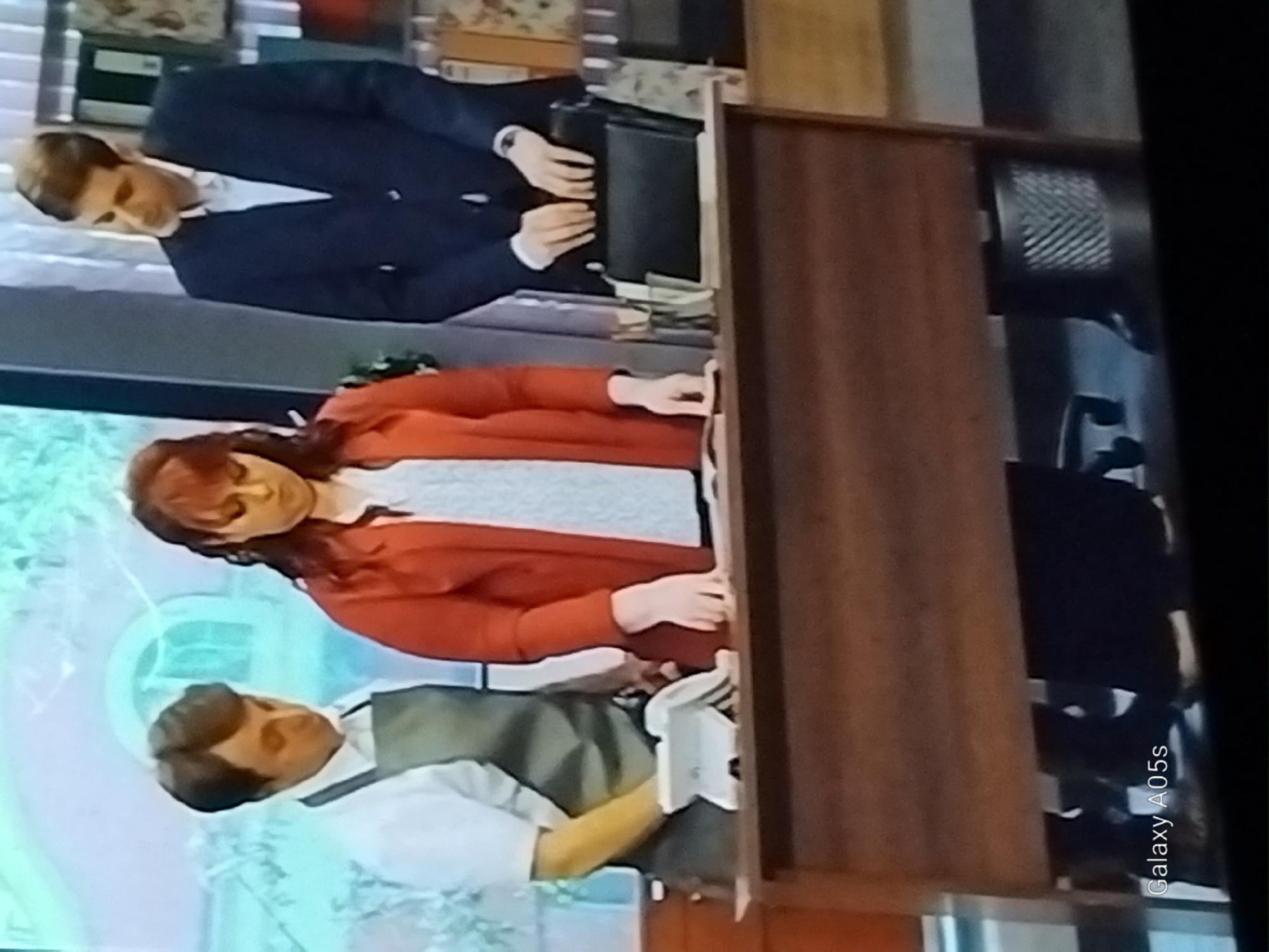 In this media file, three individuals are depicted within what appears to be an indoor setting, possibly a studio or a room with a television screen displaying a scene. The primary subjects are two men and one woman, all appearing to be adults.

The woman in the foreground is wearing a red cardigan over a white and patterned shirt. She is positioned in a way that suggests she might be interacting with the man below her, who is dressed in a white shirt and a green vest, with suspenders visible. His arm is extended, and he is holding a white object, perhaps a prop or part of the set.

The second man is at the top of the frame, wearing a dark blue suit jacket over a white shirt. He is holding a black briefcase. His gaze is directed downwards, and his hands are resting on the briefcase.

The background features elements that could be part of a television set or a room. There are shelves with objects, and a large, wooden piece of furniture, possibly a cabinet or a desk, occupies a significant portion of the right side of the frame. The lighting suggests an indoor environment, and no specific time of day or weather conditions are discernible.

There is no clear indication of a specific activity or event taking place beyond the posed nature of the individuals. The emotions of the subjects are not explicitly conveyed. A small piece of text, "Galaxy A05s," is visible vertically along the right edge of the image, likely indicating the device used to capture the media.

The provided location context, Druzhba, Russia, does not offer any specific visual cues within the image itself to confirm or deny this location.