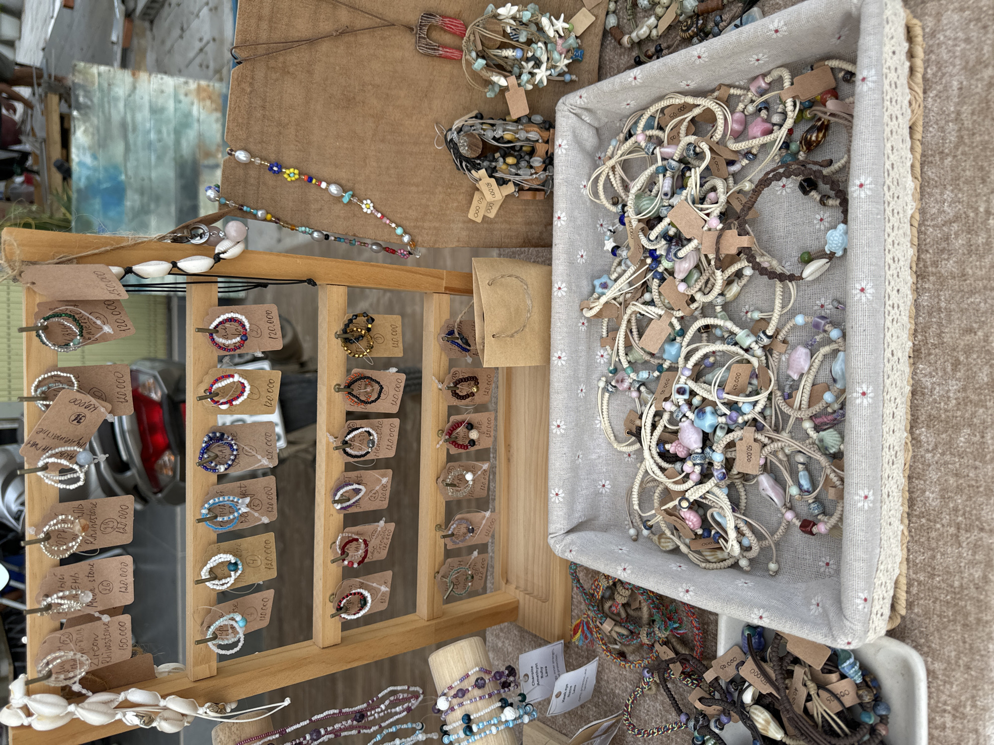 In Mũi Né, Vietnam, a market stall displays an assortment of handmade bracelets and necklaces. The stall features a wooden stand with multiple tiers holding various bracelets, each with a small paper tag indicating its price. Among the displayed items are necklaces made with beads, shells, and small charms. A woven basket filled with more bracelets is placed in front of the stand. The background shows a glimpse of a street scene with a motorcycle, suggesting an outdoor market setting. The overall ambiance is that of a typical roadside stall selling artisanal jewelry.