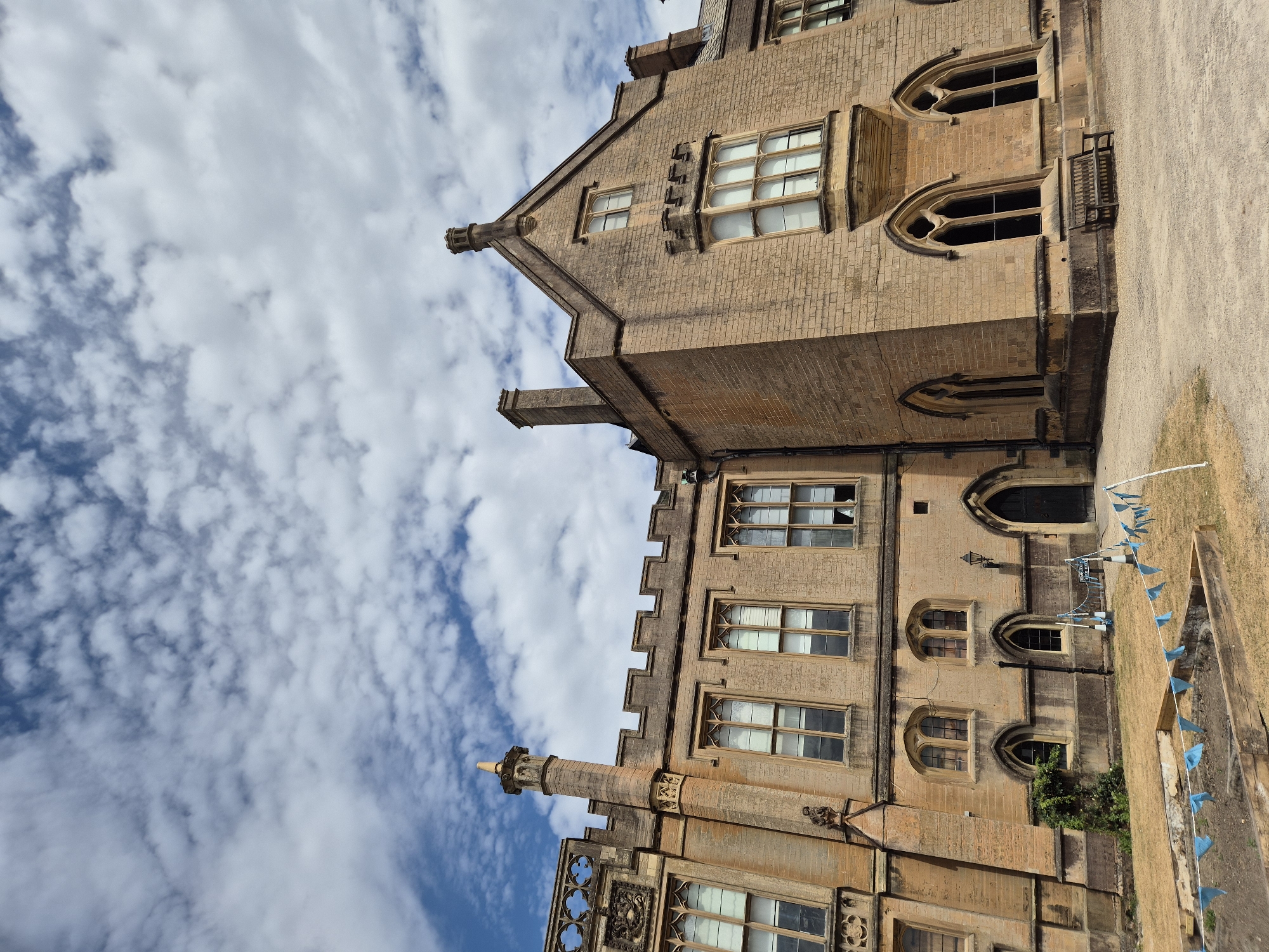 The image shows a large, stone building with a gothic architectural style. The building has multiple windows, some of which are arched. There are also chimneys and decorative elements on the roofline. The sky is blue with scattered white clouds. In the foreground, there is a patch of dry grass, a wooden bench, and some blue flags strung between poles. The building appears to be an old historical structure, possibly a school or a government building. There are no people visible in the image. The location is not specified as Nottingham, United Kingdom.