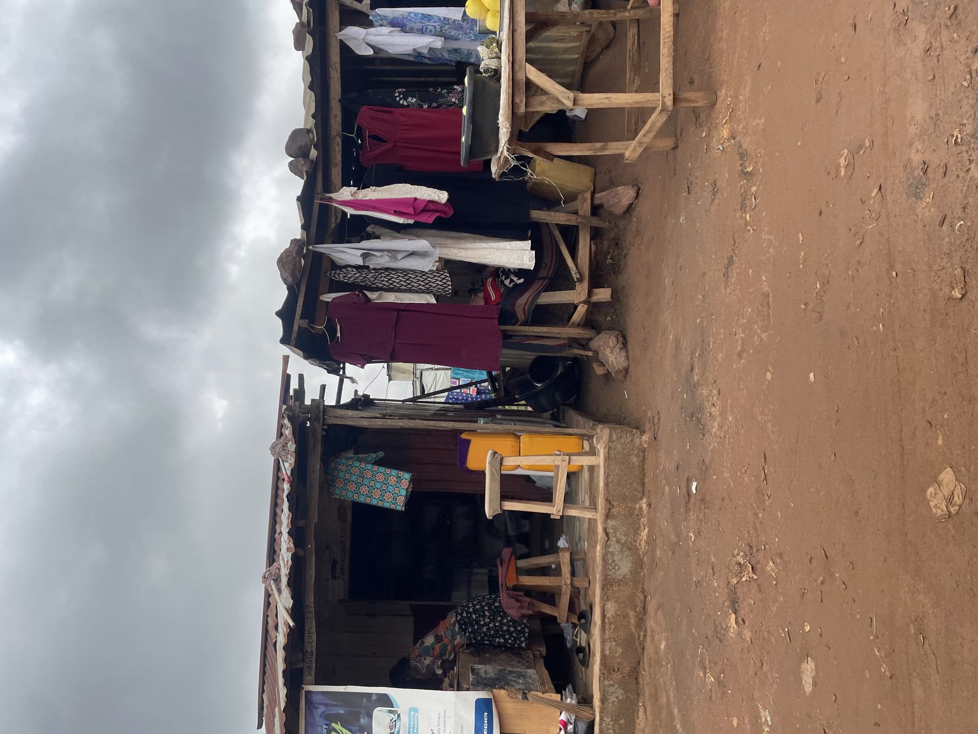 A woman stands inside a small, open-air shop in Kurunduma, Nigeria, bent over a table. The shop appears to be made of corrugated metal and wood, with a makeshift roof. Clothes are displayed on a rack outside the shop, hanging from hangers. A variety of garments are visible, including dresses and patterned fabrics. In front of the shop, there are two wooden stools and a stack of yellow containers. The ground in front of the shop is dirt, with some scattered debris. The sky is overcast.