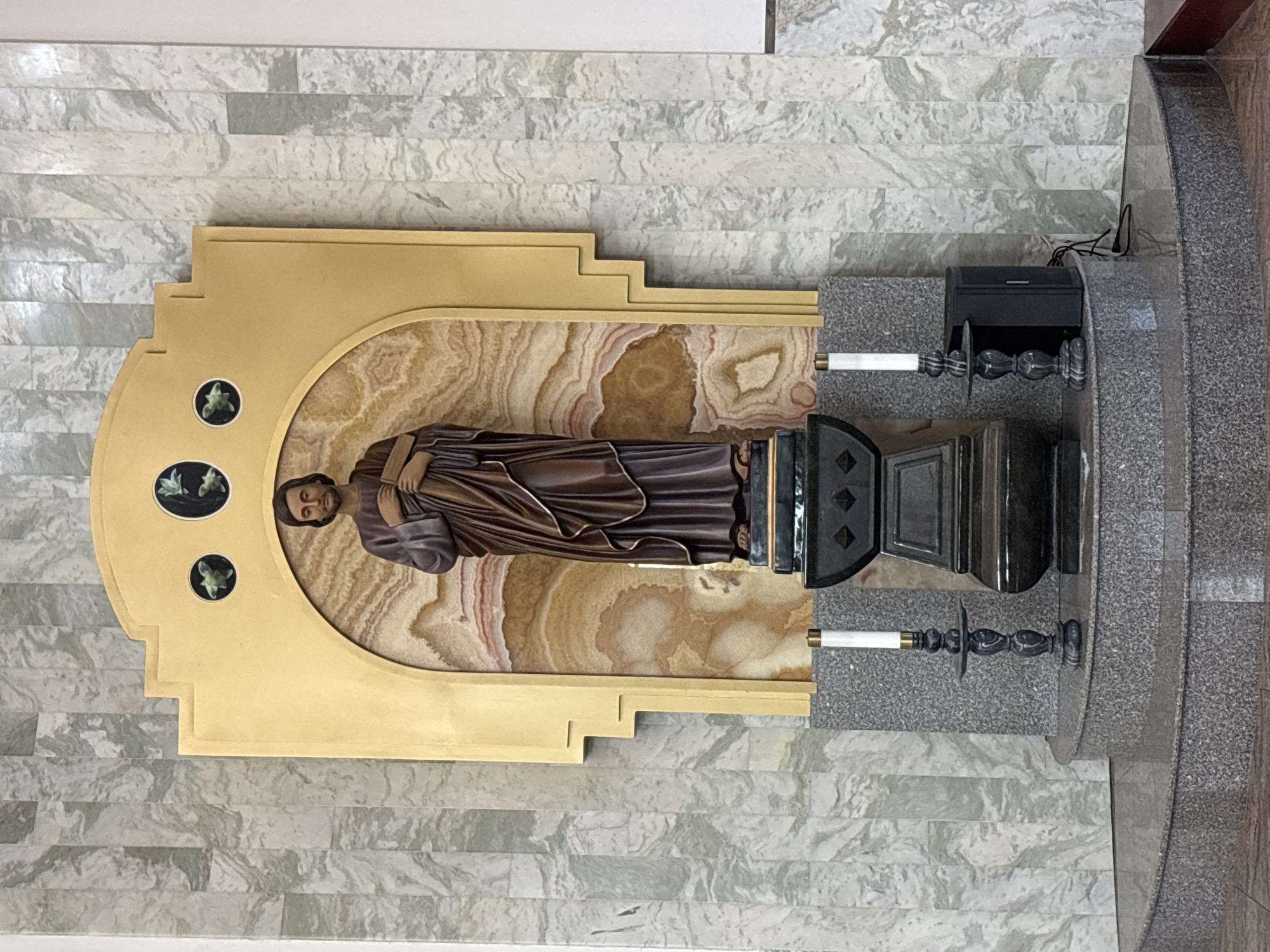 The image depicts a statue of a male figure, possibly a saint, dressed in a brown robe, standing within a golden niche against a backdrop of marbled stone. The statue is positioned on a dark pedestal with ornate carvings. Two candle holders with white candles flank the pedestal. The entire setup is situated on a multi-tiered granite base. The background wall is covered in vertically aligned, pale green and white marbled tiles. The scene appears to be inside a religious building, such as a church or chapel. There are no visible people in the image. The location is not identifiable from the provided image and therefore cannot be stated.