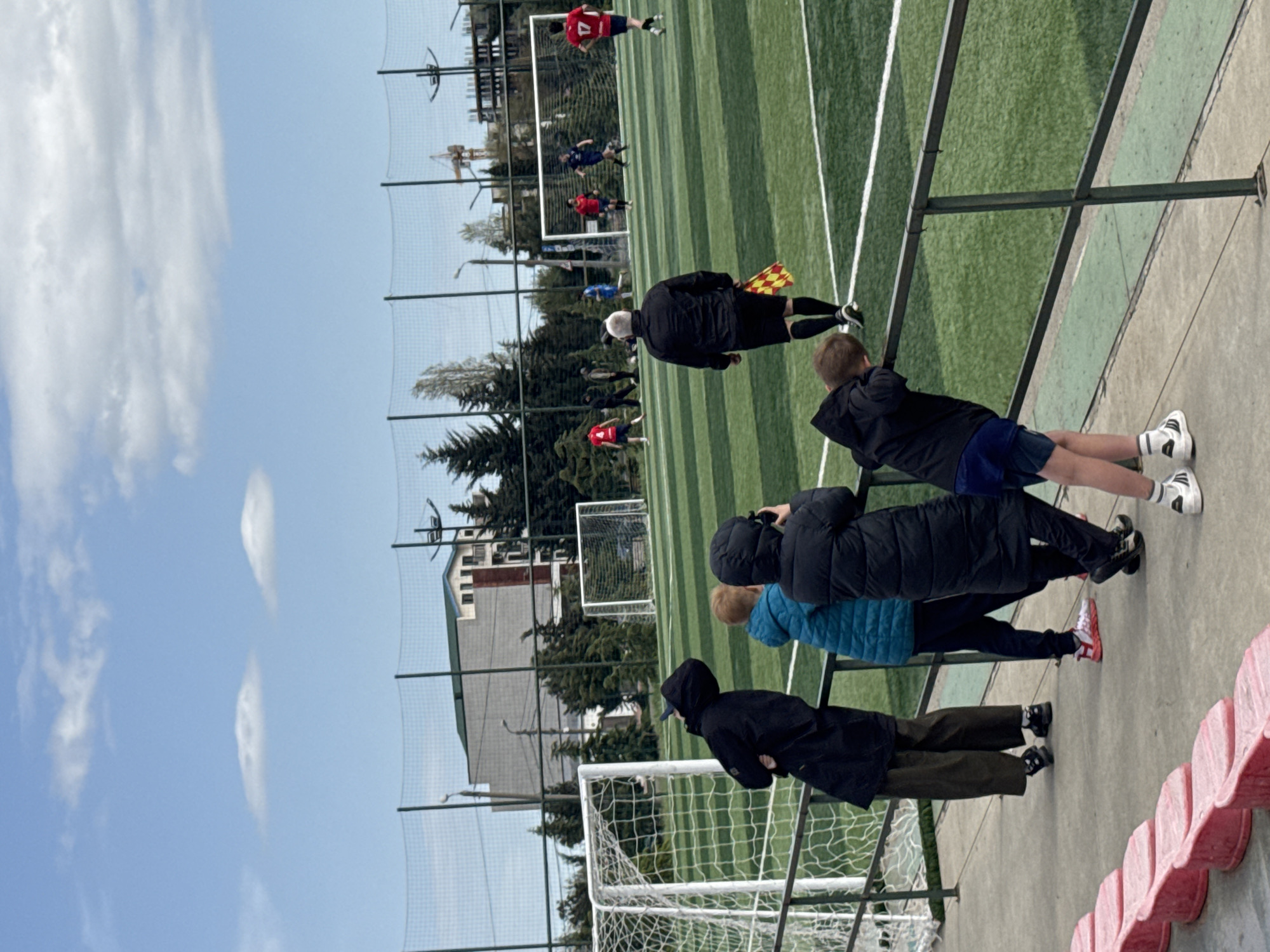 The image shows a soccer game in Tbilisi, Georgia. Several spectators, including children and an adult, are watching the game from the sidelines. One referee is visible on the field. A soccer field with green artificial turf marked with white lines is observed. Beyond the field is a net fence and some buildings and trees are present. The sky is blue with clouds.