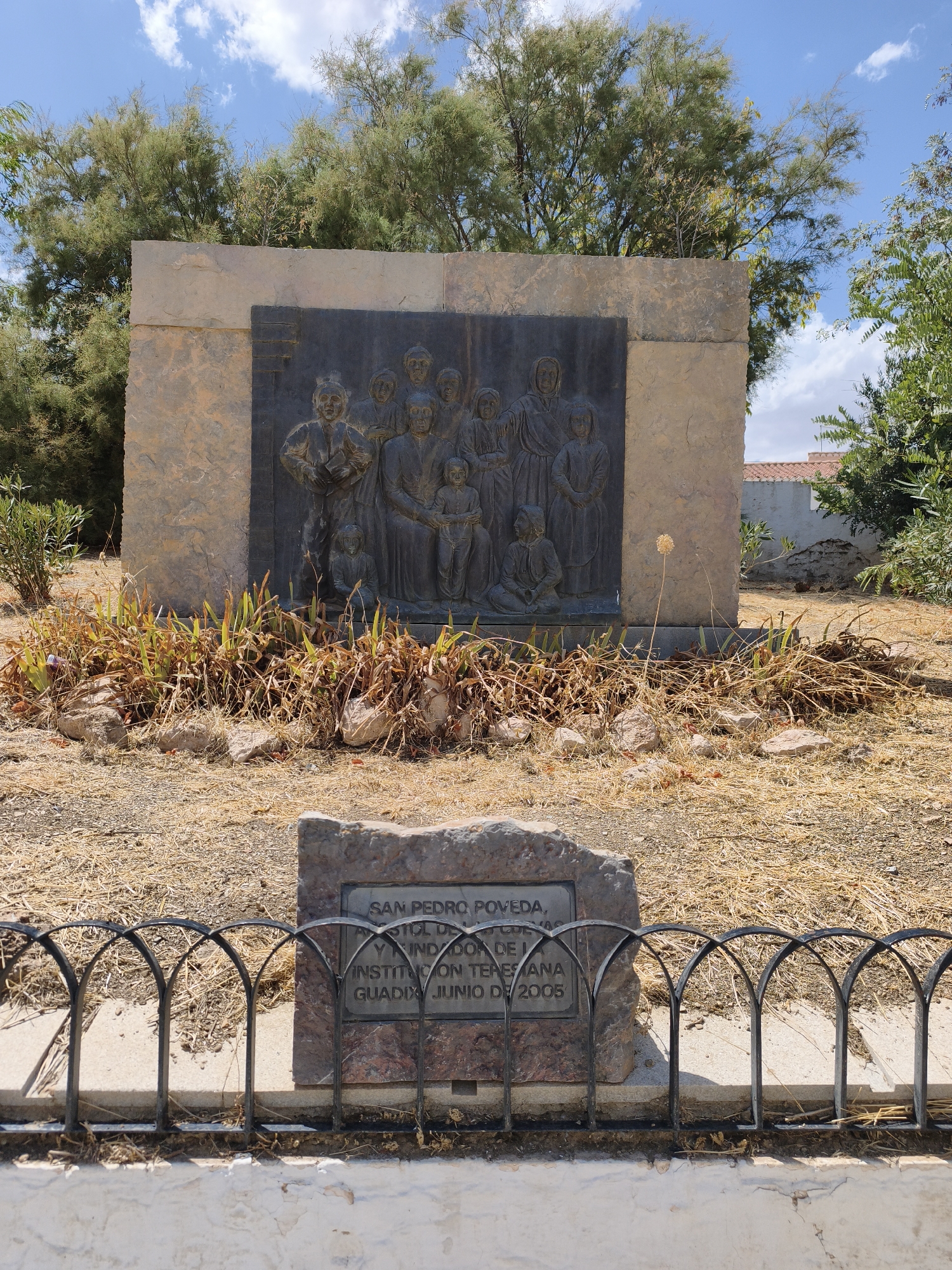 This image depicts an outdoor monument in Guadix, Spain, likely within a park or public square. The monument features a large, rectangular stone slab with a dark metal relief sculpture on its face. The sculpture portrays a group of individuals, appearing to be a family or community, depicted in a formal, posed manner. The figures are rendered in a somewhat stylized, historical or traditional style, with the central figures appearing larger and more prominent.
In front of the main monument is a smaller, rough-hewn stone plaque with an engraved inscription. This plaque is partially obscured by a decorative black metal fence, which encloses the area in front of the monument. The ground surrounding the monument is dry and covered with straw-like vegetation and scattered rocks, suggesting a warm or arid climate. Behind the monument, a line of trees with light green foliage forms a backdrop, and in the distance, a white building with a terracotta roof is visible, along with more greenery. The sky above is blue with scattered white clouds. The overall impression is one of a historical or commemorative site.