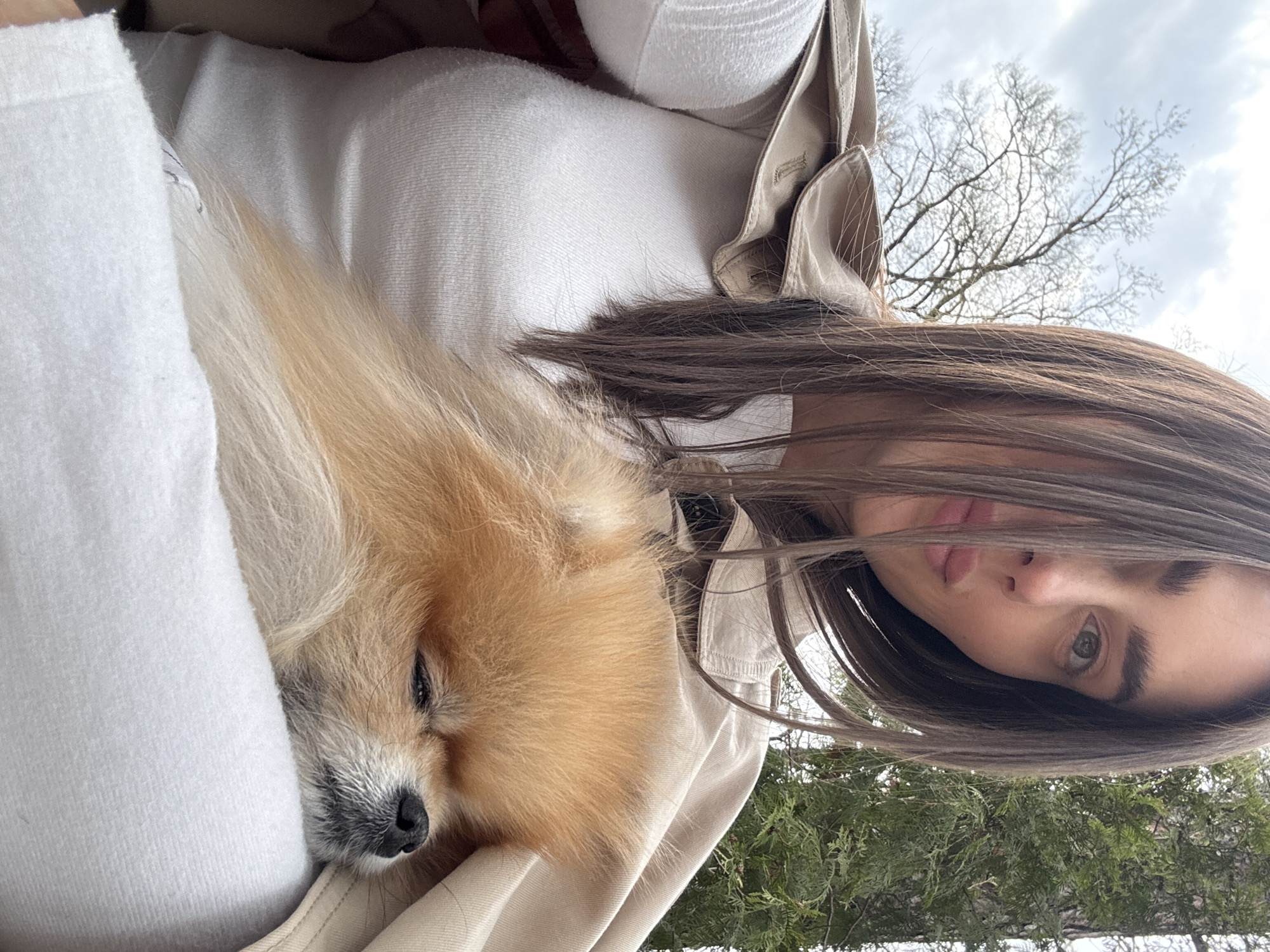 The photograph shows a person holding a Pomeranian dog. The person has long, dark hair, fair skin, and green eyes. They are wearing a white top and a beige jacket. The dog is mostly orange and is nestled in the person's arms, appearing to sleep.
Based on the surrounding environment, which includes trees and greenery, the location appears to be outdoors. The reference to Konstancin-Jeziorna, Poland, is not visually substantiated in this image. Without further information, pinpointing the specific location remains uncertain.