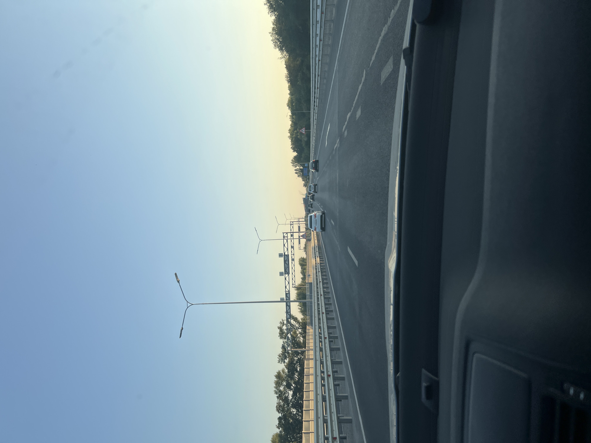 The image shows a view from inside a vehicle driving on a highway. The sky is clear and blue, transitioning to a lighter hue near the horizon. Multiple lanes of asphalt road are visible, with guardrails lining the sides. Several cars are driving on the highway in the same direction. Tall streetlights are positioned along the road. On the left side, there is a line of trees behind a metal fence. On the right side, there are also trees lining the edge of the highway. A traffic sign indicating a possible hazard or caution is visible on the right side of the road. The interior of the vehicle, including the dashboard, is visible in the foreground. The scene appears to be a typical highway drive in a suburban or rural setting, possibly during daytime hours with the sun setting.