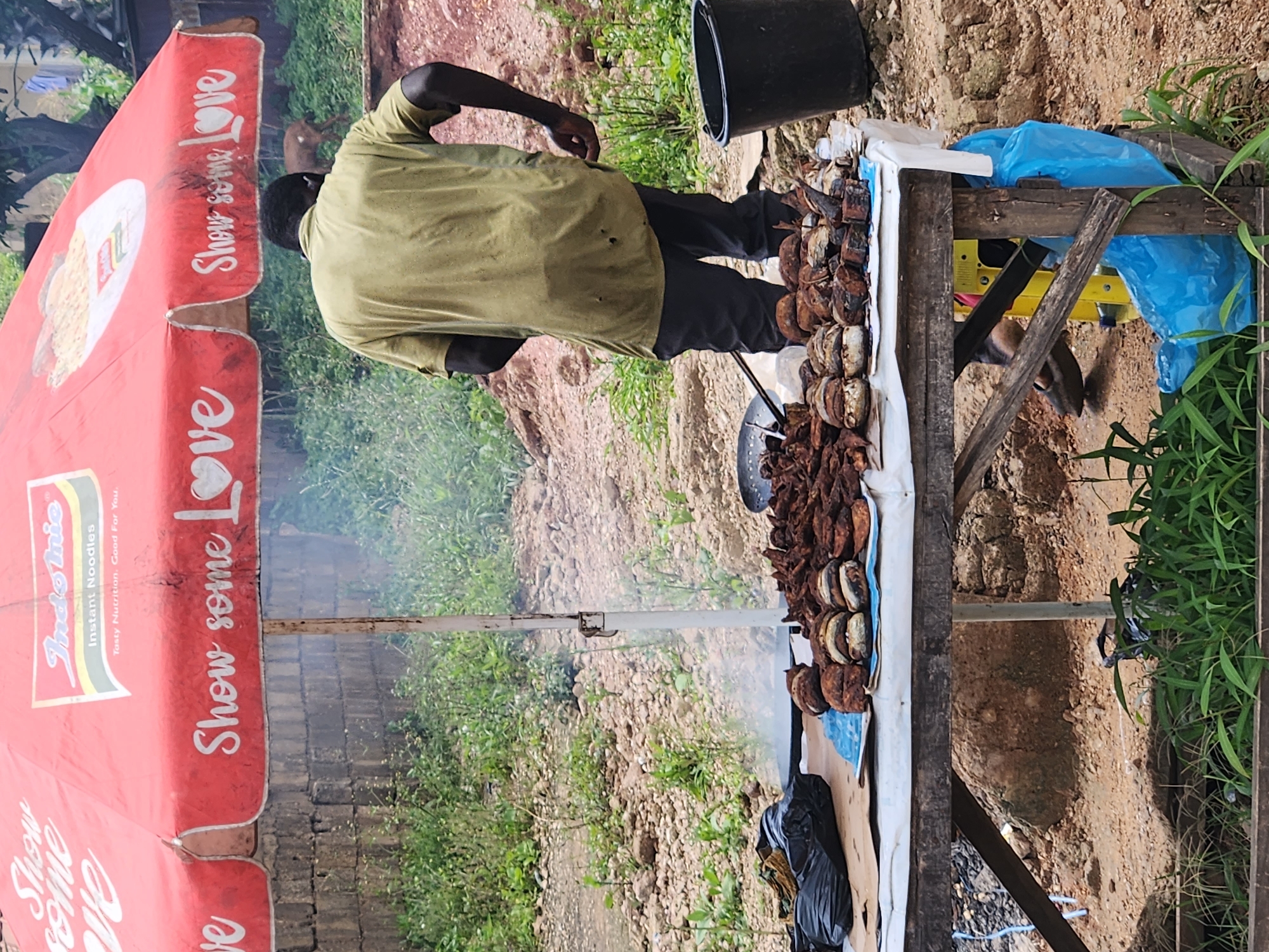An outdoor food stall under a red umbrella branded "Indomie Instant Noodles" displays grilled food items. A man in a faded olive-green t-shirt and dark pants stands behind the stall, appearing to be the vendor. He is facing away from the camera, with his right hand behind his back and his left hand near his hip. The food, a mix of what appears to be grilled fish and possibly other meats, is arranged on a white cloth atop a rustic wooden table. Several round, bun-like items with visible layers and dark, cooked exteriors are prominent among the offerings. Smoke is rising from a pan or cooking surface to the left of the food display. To the right of the stall, a black plastic bucket is visible. The ground is dirt and gravel, with patches of green vegetation. In the background, a goat can be seen near some trees, and there are structures that suggest a semi-urban or rural setting in Kabong, Nigeria. The overall impression is that of a casual street-side food vendor preparing and selling cooked items.